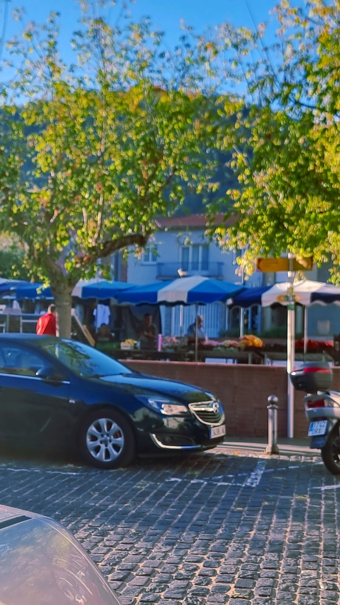 The image depicts a street scene in Sant Joan Les Fonts, Spain, with a market in the background. A dark-colored sedan is parked in the foreground on a cobblestone street. Behind the car, market stalls are set up under blue and white striped awnings, with several people visible browsing and working at the stalls. A tree with green foliage is positioned to the left, partially obscuring the market stalls and a building in the background. A portion of a motorcycle is visible on the right side of the image. The overall lighting suggests it is daytime, possibly late afternoon, with sunlight filtering through the trees.
