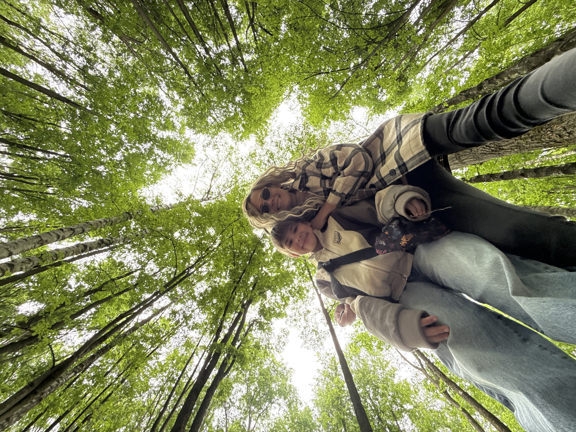 The image, taken from a low angle looking upwards, shows two people standing in a forest, with the view focused on the canopy of trees overhead. The trees are tall and slender, with dense green foliage creating a partial canopy that allows some sunlight to filter through. One person, presumably an adult, is wearing a plaid shirt, black pants, and sunglasses, with long blonde hair. The other person, a child, is wearing a light-colored jacket. Both are smiling. Rusanovo, Russia is not confirmed.
