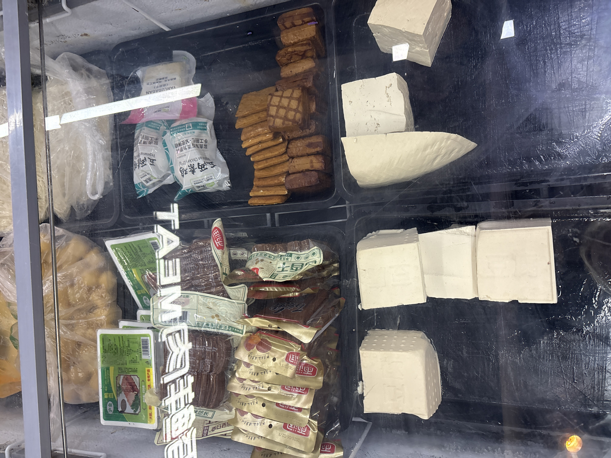 In the city of Minhang District, China, a frozen food display case contains an assortment of products. The display case is made of clear glass, allowing visibility into its contents. Inside, various types of tofu and packaged vegetarian meat substitutes are visible.
In the top section of the display, there are large plastic bags filled with what appears to be frozen produce or dough. Below this, a variety of vacuum-sealed packages of vegetarian meat products are arranged. These packages are in different shapes and sizes, with some resembling slices of meat and others in larger chunks. Many of these packages have colorful labels with Chinese text.
In the bottom section of the display case, several blocks of tofu are arranged. Some blocks are large and rectangular, while others are cut into smaller pieces. The tofu appears to be fresh and has a light, off-white color. Some of the tofu blocks have textured surfaces, with visible imprints of characters. The display case itself is well-lit, and condensation is visible on the glass, indicating a cold environment. The overall scene is typical of a frozen food section in a grocery store or market.