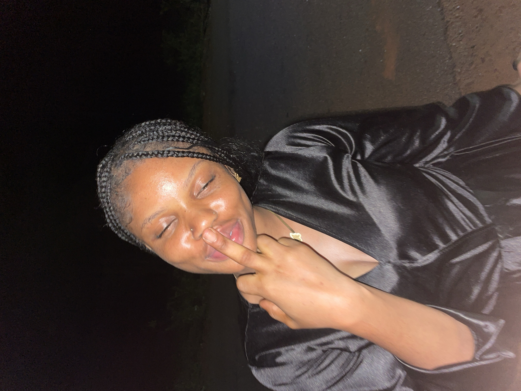 A close-up image, oriented roughly 90 degrees clockwise, captures a young Black woman in a playful moment at night, likely in Ogbe, Nigeria. She is looking towards the viewer with her eyes closed and a gentle smile, pressing her right index finger to her nose in a humorous gesture.
The woman has braided hair, styled in neat, small cornrows or box braids. Her skin appears well-hydrated and luminous, with distinct highlights visible on her forehead, cheekbones, and nose. She has a small piercing on her left nostril. She is wearing a black garment, possibly made of satin or silk, which has a lustrous sheen under the dim lighting. A delicate golden necklace with a small, light-colored charm hangs around her neck.
The background is extremely dark and indistinct, indicating an outdoor setting at nighttime with minimal ambient light. Faint, dark shapes in the background suggest distant foliage or ground textures. The primary light source appears to be directed at the subject, illuminating her face and upper body and highlighting the texture of her garment. Her expression suggests amusement and a lighthearted, mischievous mood. There is no visible text in the image.