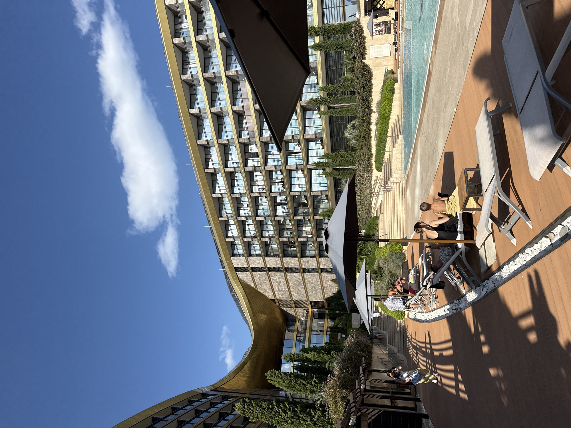 This image depicts a resort area with a modern architectural design, possibly located in the city of Simeiz, Ukraine. In the foreground, several empty lounge chairs are arranged on a wooden deck. To the right of the lounge chairs, a swimming pool is visible, with a few people relaxing by its edge under umbrellas. In the background, a large, multi-story building with numerous balconies curves around the property. The building's facade is a combination of textured stone and glass, topped with a gold-colored roof. Lush greenery and trees are planted around the building and pool area, suggesting a well-maintained outdoor space. The sky is blue with scattered white clouds, indicating fair weather. The overall scene suggests a leisurely atmosphere at a high-end vacation destination.