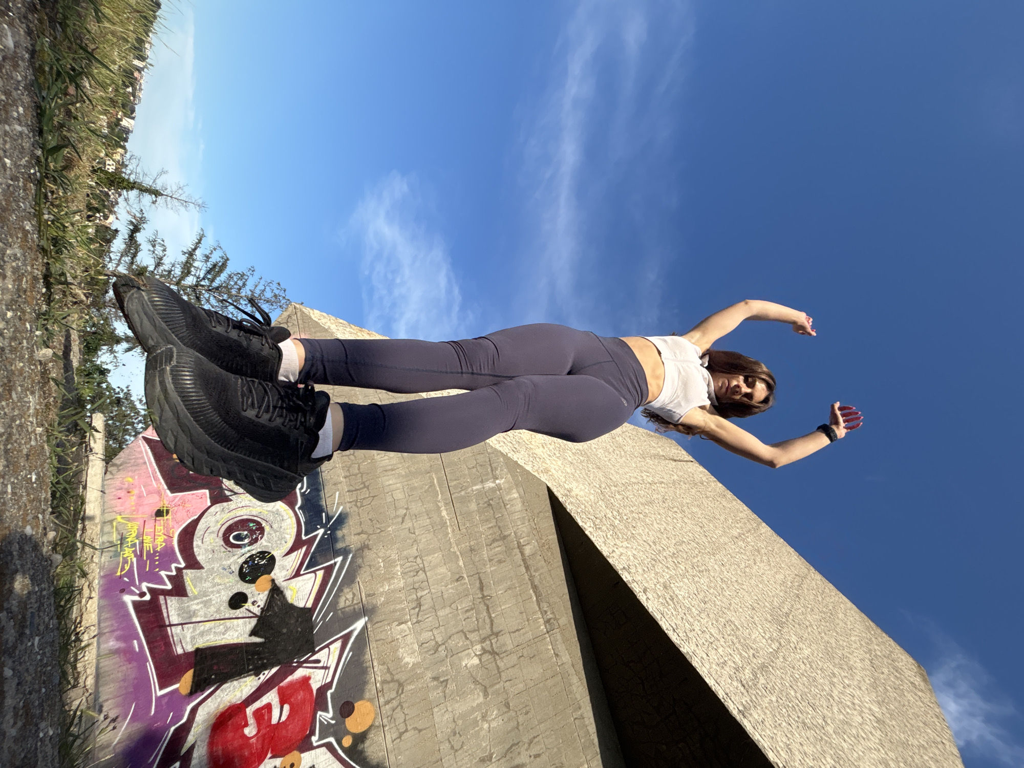 A young woman in athletic wear, including a white sports bra and dark leggings, is captured mid-air, jumping with her arms raised. She is wearing black sneakers and white socks. She is positioned in front of a concrete structure, which appears to be part of a bridge or overpass, adorned with colorful graffiti. The background features a clear blue sky with wispy clouds, and some green vegetation and a distant cityscape can be seen at the bottom left of the frame. The scene suggests an outdoor urban environment, possibly in the city of Varna, Bulgaria, given the context of the query. The low-angle shot emphasizes the woman's dynamic movement against the expansive sky and the graffiti-covered concrete.