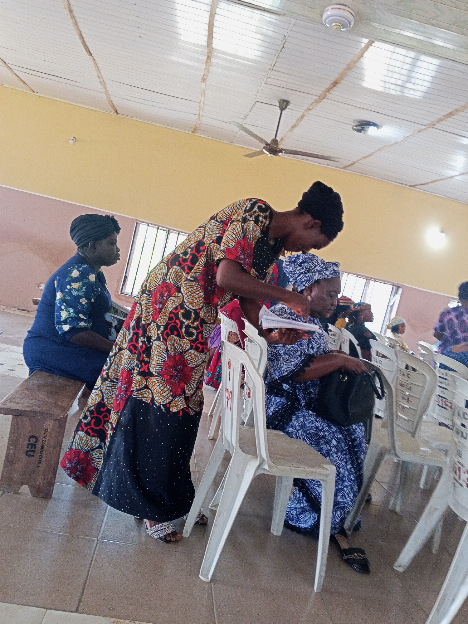 The image shows three women in a room in Zango-ndaji, Nigeria. The woman in the center, wearing a black, red, and yellow patterned gown, stands and is handing papers to a seated woman wearing a blue and white patterned outfit. The seated woman has a black purse on her lap. To the left, another woman in a blue floral dress sits on a wooden bench. The room has a light yellow upper wall and a white ceiling with a ceiling fan and recessed lighting. Rows of white plastic chairs are present. The floor is tiled. The bench has the label "JACS ZANGO CITY 1 CEU."