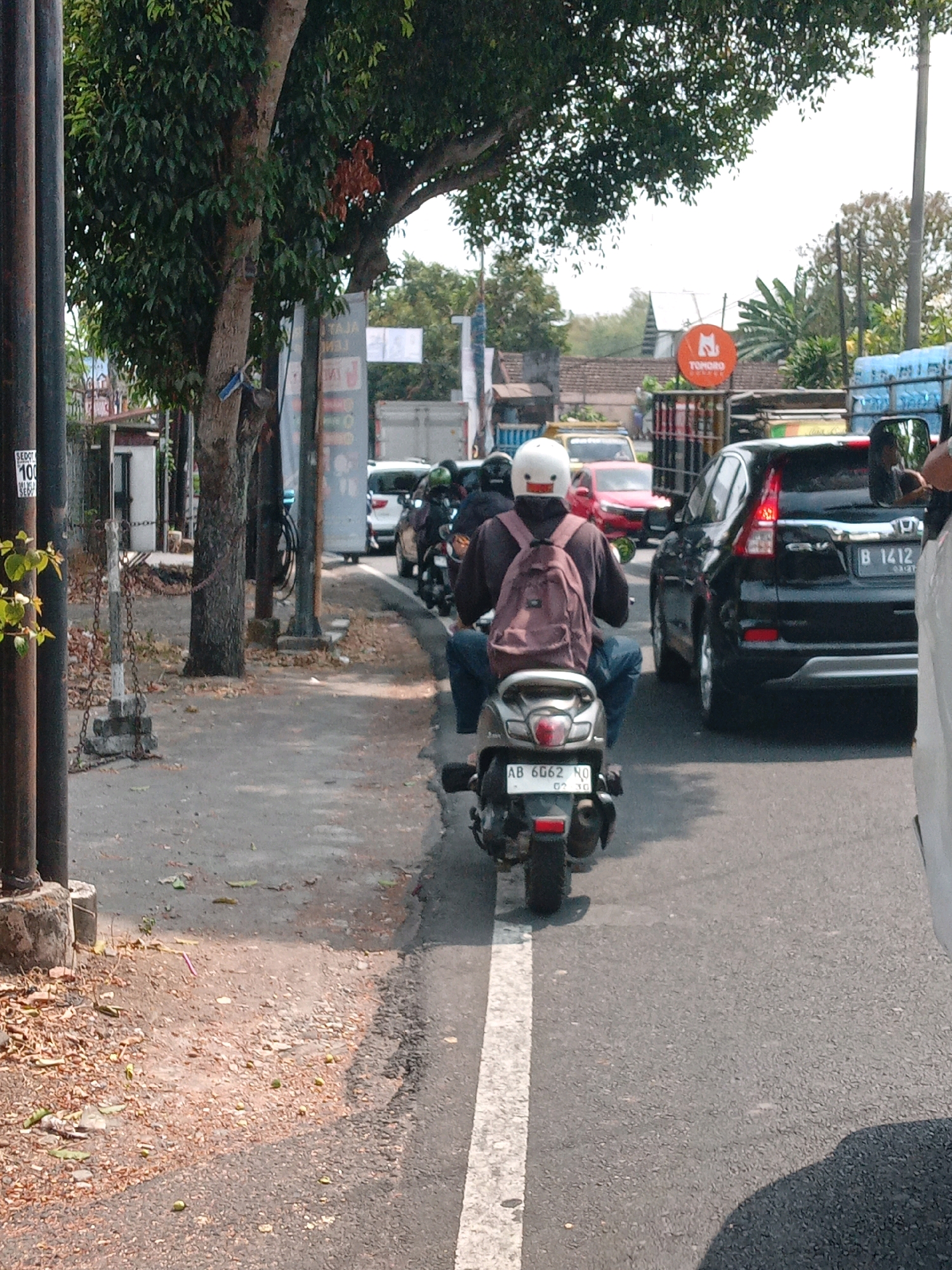 The image depicts a street scene in Ngaglik, Indonesia, with a focus on traffic and pedestrians. In the foreground, a person wearing a white helmet with a unique design and a brown backpack is riding a scooter. The scooter has a license plate that reads "AB 6062 NO". They are traveling on a paved road marked with a white line. Several other vehicles, including cars and motorcycles, are visible in the background, indicating moderate traffic. A large tree with dense green foliage shades a portion of the scene. On the left side of the image, a sidewalk area is visible with some fallen leaves and a decorative chain barrier. Buildings and signs are present further in the background, suggesting an urban or semi-urban environment. The overall lighting suggests it is daytime, with bright sunlight casting shadows.