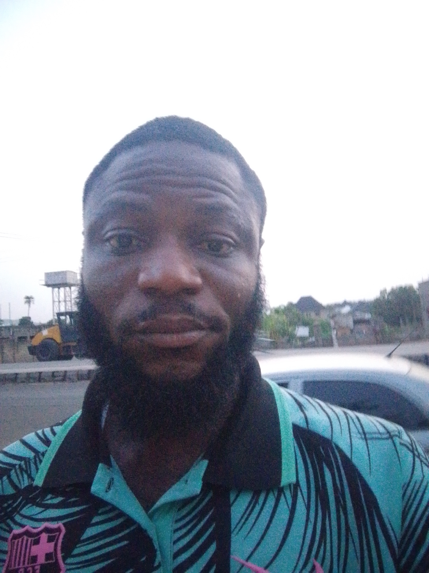 A dark-skinned man with a beard is taking a selfie. He is wearing a turquoise and black patterned polo shirt with a Barcelona FC emblem on the lower left. A car is partially visible behind him. In the background, there is a water tower on top of scaffolding, a bulldozer, and some buildings. The location is potentially Lokoja, Nigeria.