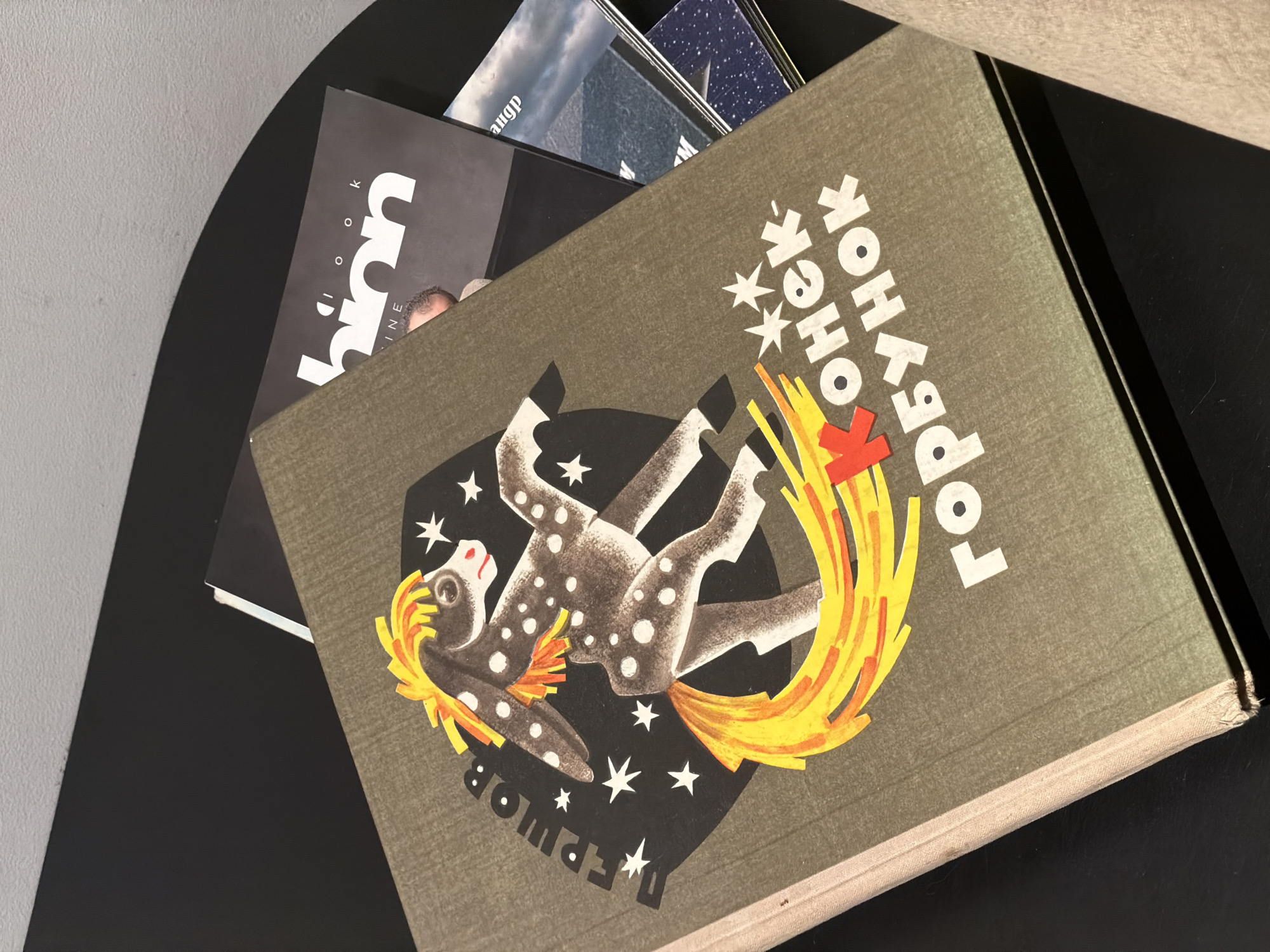 The image shows a close-up of a stack of books on a black table, likely indoors. The topmost book has a dark green cover with a graphic of a stylized, spotted horse leaping through flames, surrounded by white stars. Cyrillic text is visible on the cover. The book is likely a copy of a children's book called "Konyok-Gorbunok," which translates to "The Humpbacked Horse." It is a classic Russian fairy tale written in verse by Pyotr Yershov. The Cyrillic characters "ГЕРШОВ" are visible, suggesting the author's name. The overall scene doesn't provide explicit information to indicate Moscow or its specific location, but the book itself is representative of Russian culture.