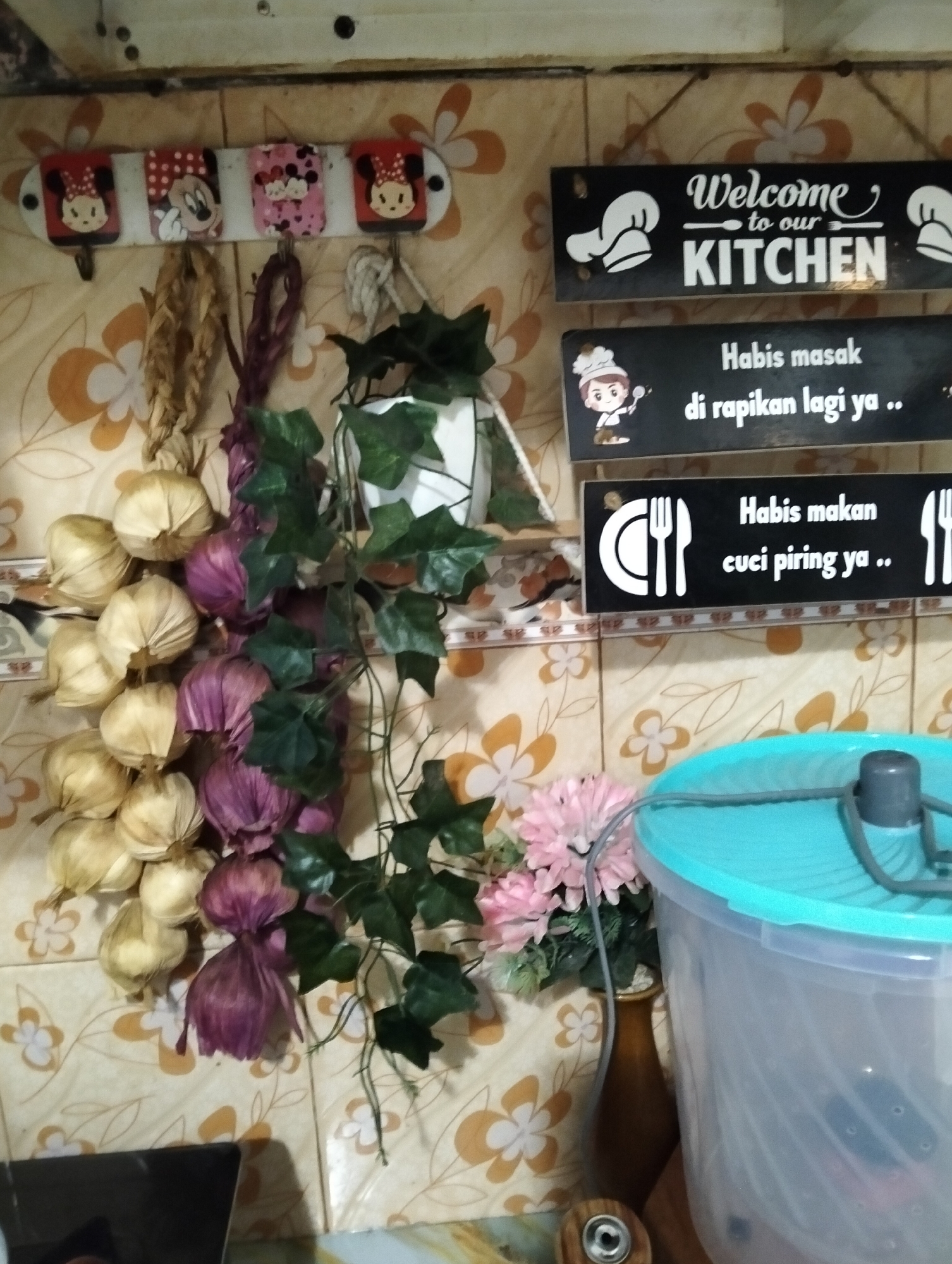 In a kitchen, a string of dried onions and garlic hangs from a white rack with four Mickey Mouse-themed decorative hooks. Above the hanging produce, two black signs are mounted on a tiled wall. The upper sign reads "Welcome to our KITCHEN" in white text with chef hat graphics. The lower sign has a cartoon chef graphic and the Indonesian text "Habis masak di rapikan lagi ya.." which translates to "After cooking, tidy up again..". To the right of the signs, a large, transparent plastic container with a turquoise lid is partially visible. In front of the container sits a small brown vase holding a bouquet of pink artificial flowers and greenery. The wall behind these items is covered in light-colored tiles with an orange floral pattern.