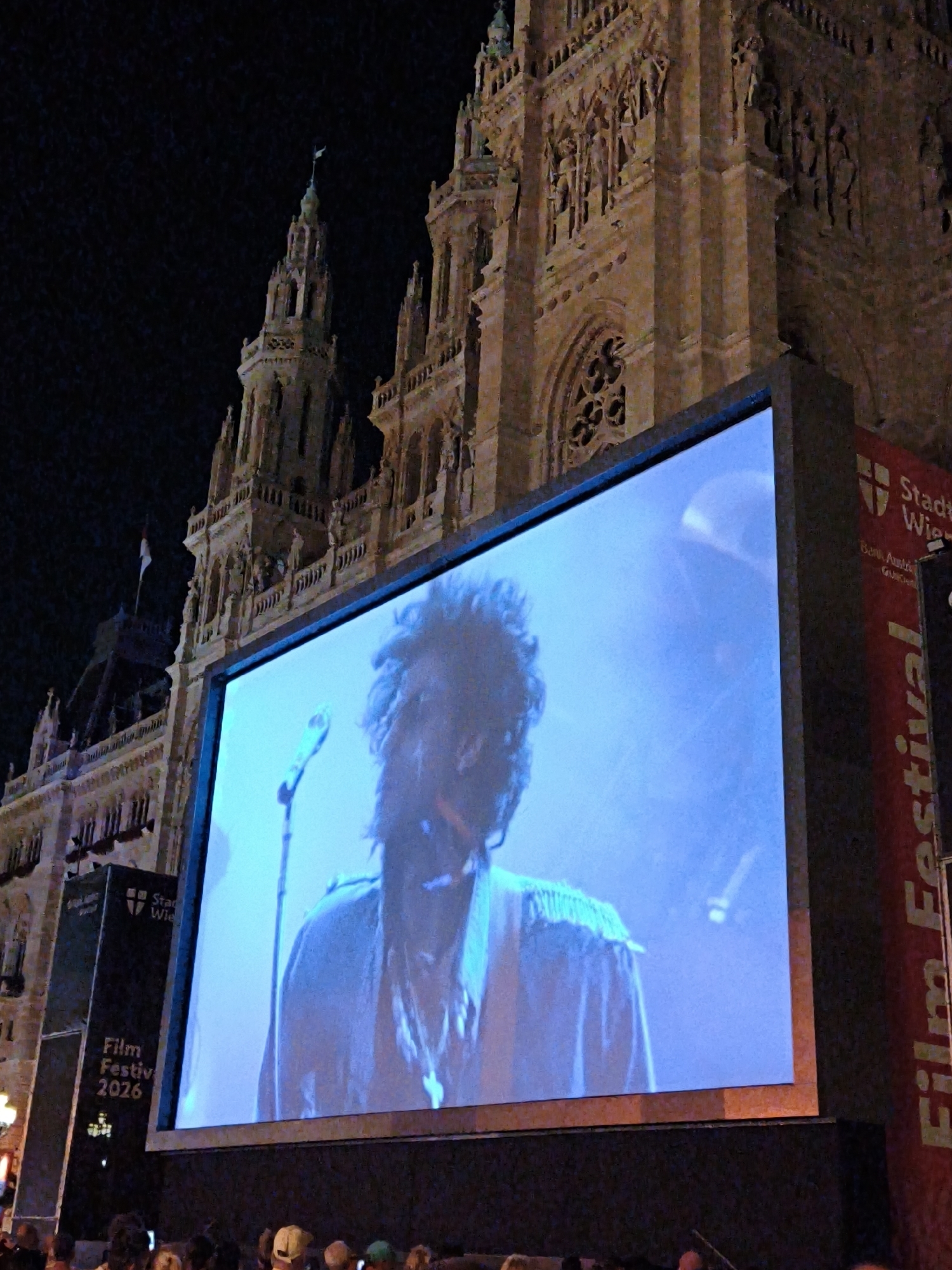 The image depicts an outdoor film screening at night in front of the Rathaus (City Hall) in Vienna, Austria. A large screen displays a close-up of a musician, likely a performer, with a microphone visible. The musician is wearing a light-colored shirt with fringe detail on the shoulders. The Rathaus, a neo-Gothic architectural masterpiece, is illuminated and forms the backdrop for the event. A vertical banner on the right side of the screen reads "Film Festival" and "Stadt Wien" (City of Vienna). The crowd in the foreground is silhouetted against the screen, suggesting they are watching the performance. The scene suggests a public event taking place in a prominent urban location.