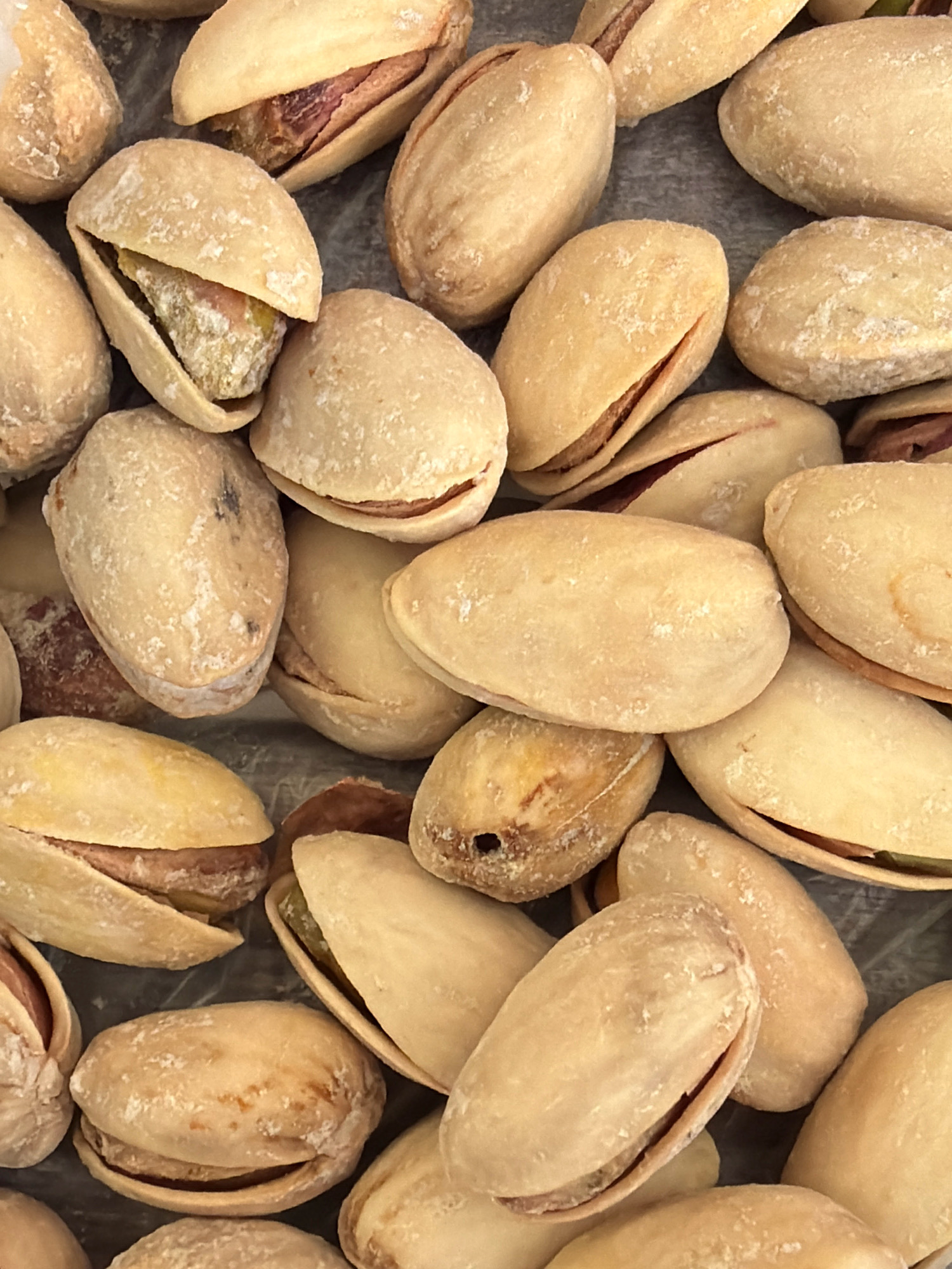 A close-up, overhead shot displays a multitude of roasted and salted pistachios, piled together. The nuts vary in their degree of openness, revealing the reddish-brown skin and pale green kernels within. Some pistachios are dusted with white salt crystals, indicating they are a salted variety. The textured, dark grey surface beneath the pistachios provides a subtle contrast to the warm, neutral tones of the nuts. The image captures the appetizing texture and appearance of the pistachios, suitable for a food-related context. There are no discernible elements in the image that suggest a location in Kyiv, Ukraine, or any specific urban environment.