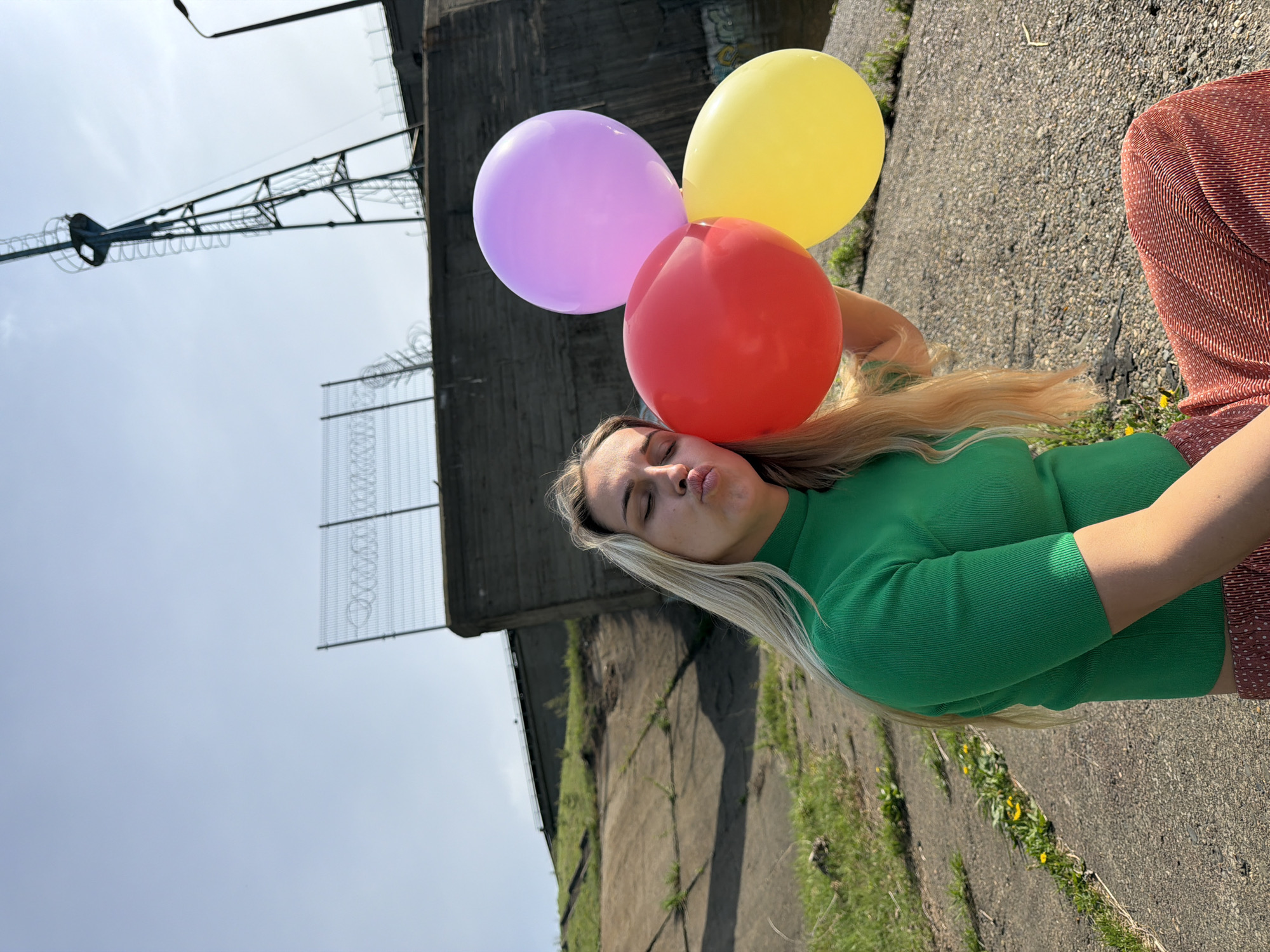 A young, blonde-haired woman is posing with three balloons (red, yellow, and purple) against a concrete structure with a metal lattice on top. She is wearing a green top and a reddish-brown patterned skirt. The woman is making a kissing face. The setting appears to be outdoors, likely near the city of Chaikovsky, Russia. Some grass and vegetation are visible near the bottom of the image along the concrete.
