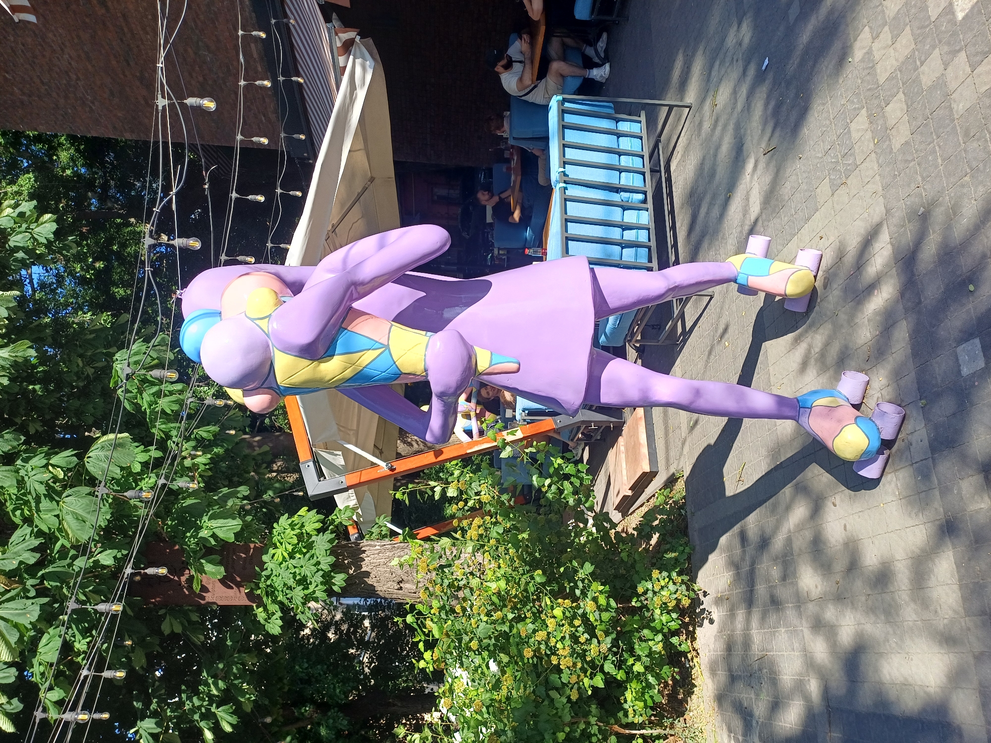 This media file features a large, abstract purple statue of a figure with roller skates on its feet, set in an outdoor urban environment. The statue's body is composed of large, rounded shapes and it wears a garment with a colorful, geometric pattern in yellow, blue, and light pink. In the background, there are people sitting at tables under an awning and a blue tiered seating structure. String lights with visible bulbs are strung across the scene. The ground is paved with grey tiles and surrounded by greenery and trees. The lighting suggests it is daytime, with strong shadows cast by the statue and other objects, indicating sunny weather. The presence of outdoor seating and people suggests a public space, possibly a cafe or recreational area in Odesa, Ukraine. There is no visible text in the image.