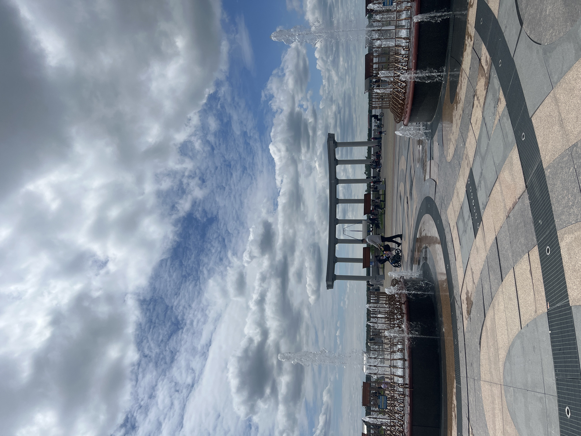 This full-frame image depicts an outdoor public space on a partly cloudy day, identified as the Amur Cliff (Utyos) viewing platform in Khabarovsk, Russia. The sky above shows a mix of large, dense grey clouds and patches of clear blue sky with smaller, scattered white clouds.
In the foreground, two circular fountains are prominently featured, each actively spraying streams of water upwards. These fountains are encased by decorative metal railings and set into a paved surface. The paving exhibits a geometric pattern of concentric arcs and radial lines, composed of dark grey, light grey, beige, and reddish-brown tiles, interspersed with dark metal drainage grates.
The midground reveals a wide promenade or plaza where several individuals and small groups are present. One person, wearing a light-colored long-sleeved shirt and dark trousers, is seen pushing a dark baby stroller from the right side of the frame towards the left. Other figures are either walking or seated on benches positioned along the promenade.
In the background, a distinctive architectural structure is visible. It consists of multiple tall, light-colored cylindrical columns supporting a flat entablature, forming an open, colonnaded archway. Beyond this structure, a broad body of water, consistent with the Amur River, extends towards a distant, slightly hazy horizon.