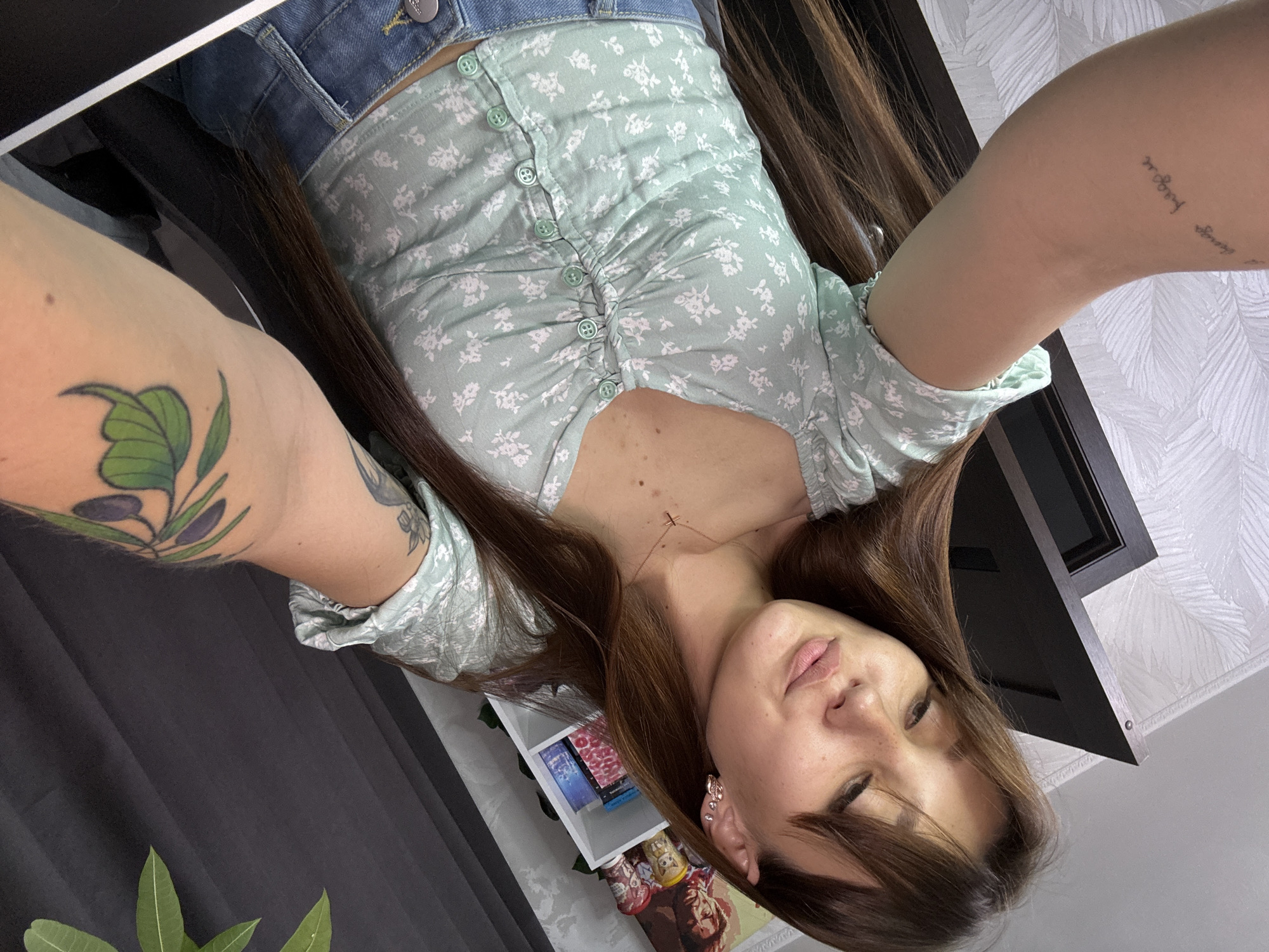 The image shows a young woman with long brown hair and bangs, taking a selfie. She is wearing a light green floral print top with buttons and blue jeans. She has freckles on her face and is wearing a small cross necklace and earrings. There is a tattoo of an olive branch on her right arm and a script tattoo on her left arm. In the background, there is a white shelf with books and decorations, a dark door, and a dark curtain. The scene appears to be indoors, likely in a residential setting. There is no indication of the specific location within Novokuznetsk, Russia.