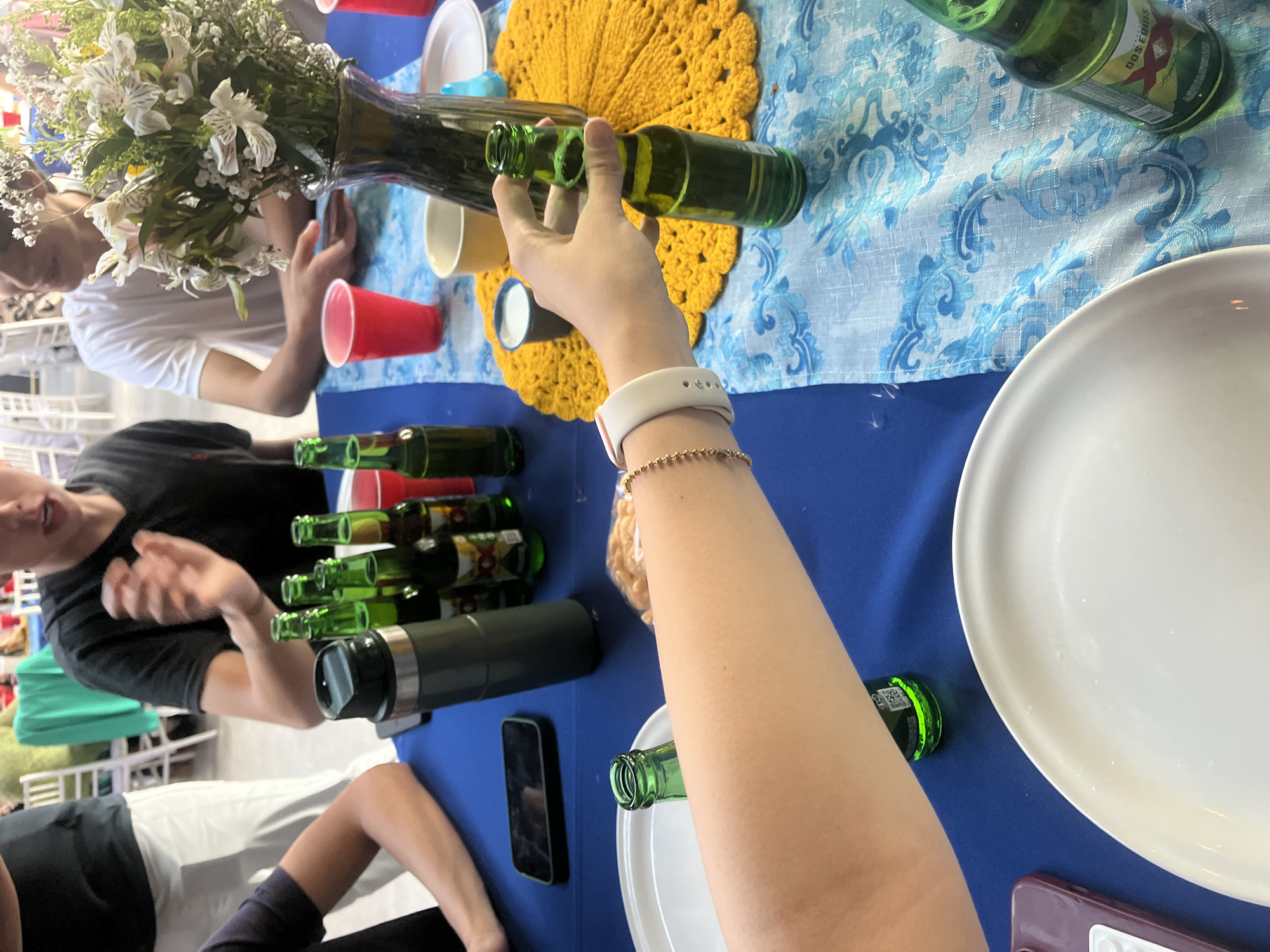 The image captures a gathering of people around a table covered with a blue tablecloth. Several green beer bottles are scattered across the table alongside red and yellow plastic cups, white plates, and personal electronic devices like phones. A vase with flowers sits prominently on the table. One person is holding a beer bottle, while others are seated, seemingly engaged in conversation. The setting appears to be a casual social event. The specific location in Puebla, Mexico, cannot be definitively determined from the provided image alone.
