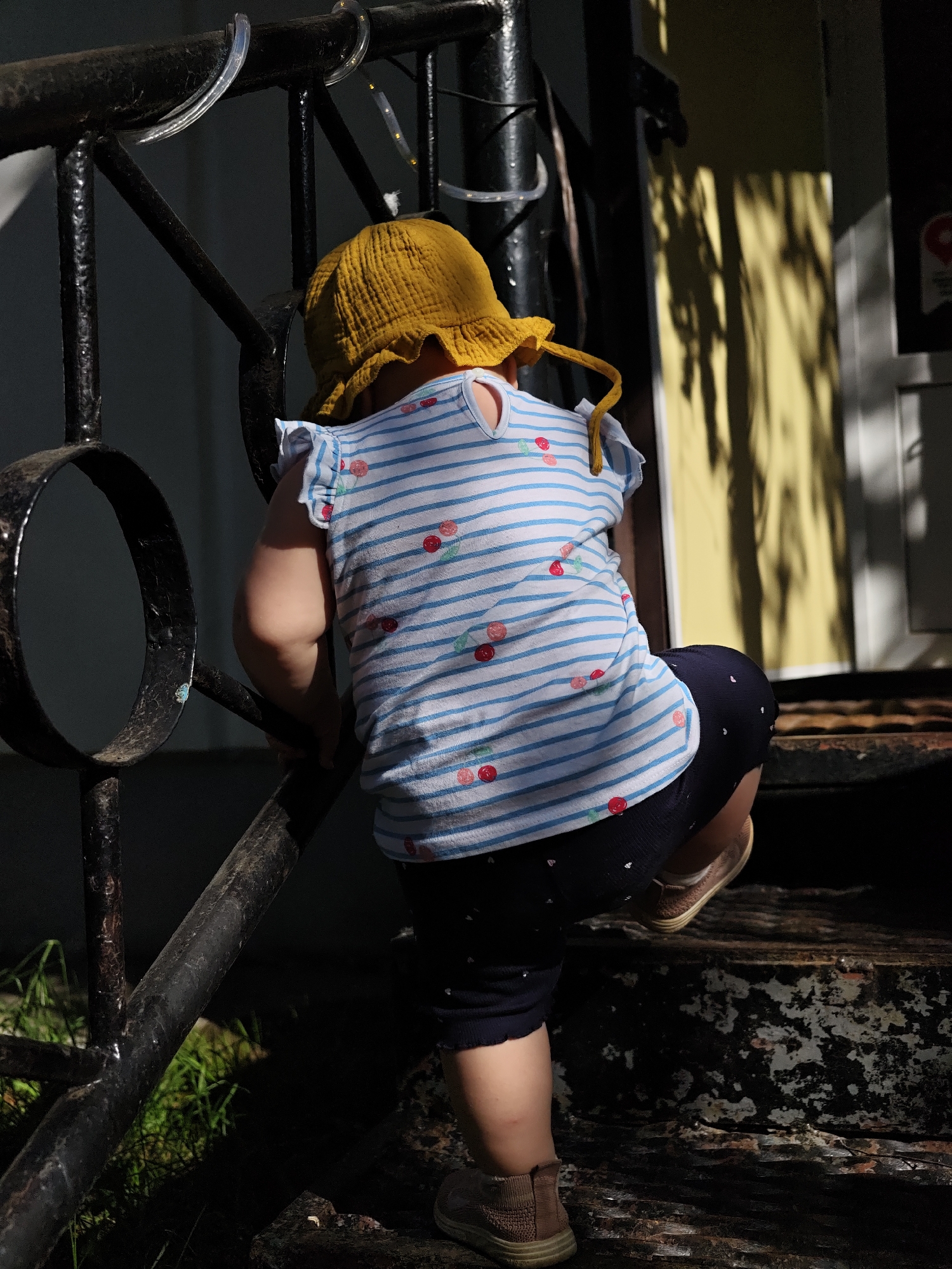 A small child wearing a yellow hat, a white and blue striped shirt with red cherries, and navy shorts with white hearts, is climbing a set of outdoor stairs with a black metal railing. The stairs are weathered and show signs of wear. The background includes a house with yellow siding. The location is Chaikovsky, Russia.