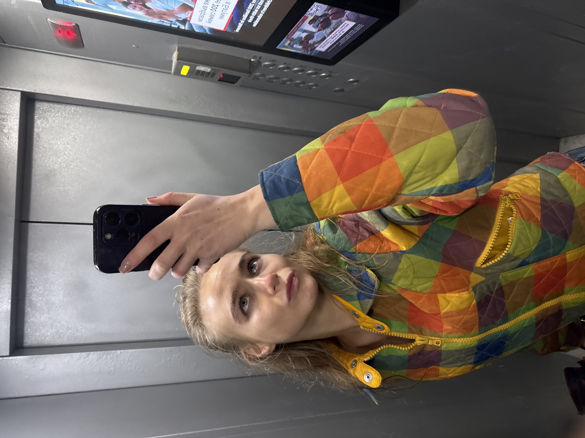 A woman is taking a selfie in an elevator. The elevator has a grey interior and features a digital display screen and a control panel. The woman is wearing a multi-colored quilted jacket. She is holding a black smartphone. The city and exact location are Unknown, Unknown.