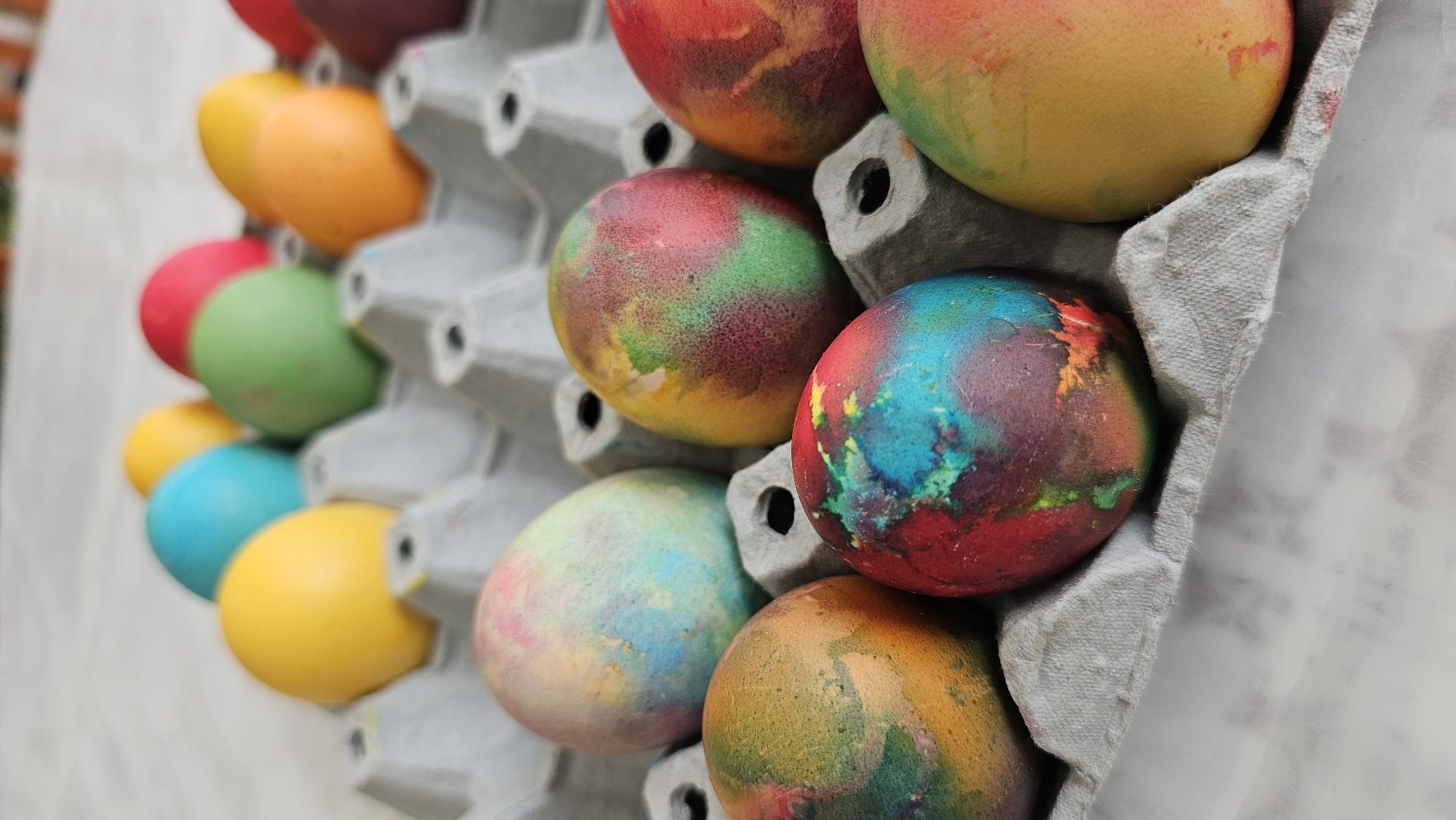 The image shows a carton of dyed Easter eggs. The eggs are varying colors including solid yellow, orange, red, green, and blue, and some with mixed colors and marbled patterns. The egg carton is gray. The background is blurry and appears to be a white surface. There are no people visible in the image. The location is not discernible from the image.