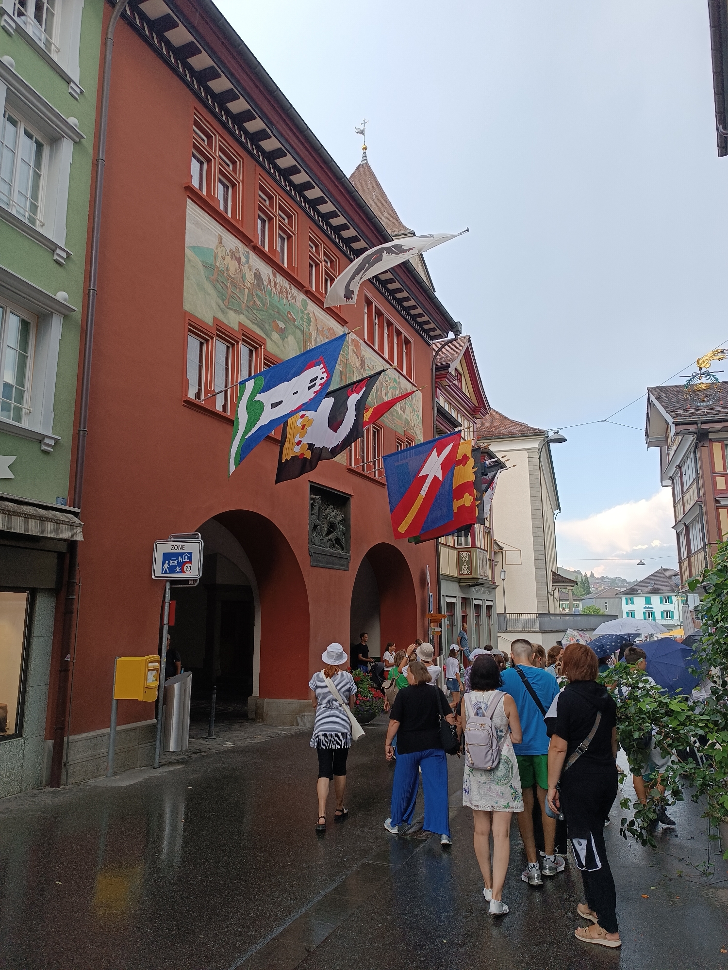In Appenzell, Switzerland, a street scene unfolds with a parade of people walking on a wet cobblestone street. The architecture features a prominent red building with arches, adorned with several colorful flags flying from its facade. Adjacent to this building, a lighter green building stands, its windows reflecting the overcast sky. The group of people is diverse, with individuals of various ages and genders, some carrying umbrellas, suggesting recent rain. They are walking in the same direction, away from the viewer, on a sidewalk that appears to have been recently washed. The overall atmosphere is that of a town event or a casual stroll through a historic European town.