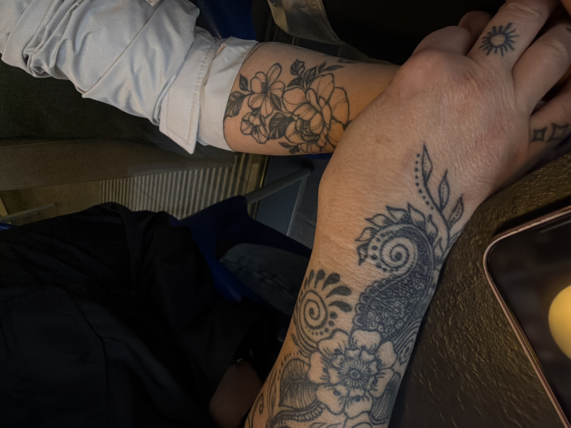 The image shows two arms with tattoos. One arm, wearing a light-colored, cuffed jacket, has a floral tattoo on the forearm. The other arm, which is bare, has a larger, more intricate tattoo that includes floral and paisley designs, extending from the wrist up the arm. There is also a tattoo of a sun on one of the fingers and small diamonds. A portion of a smartphone is visible, and fabric from clothing is also visible. The location in the city of Unknown, Unknown is not discernable from the image.
