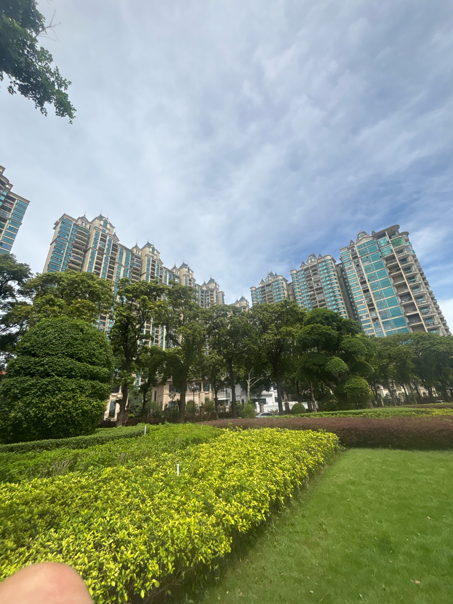 This image captures a view of high-rise apartment buildings in the city of Nanhai District, China. In the foreground, a lush green lawn is bordered by neatly trimmed hedges with bright yellow-green foliage. Behind the hedges, a band of darker, reddish-brown bushes forms a contrasting line. Mature trees with dense green canopies are scattered throughout the scene, providing shade and separating the landscaped area from the buildings. The apartment buildings themselves are tall and modern, with many floors and balconies. Their facades feature a combination of light-colored material and turquoise glass windows, and they are topped with decorative architectural elements. The sky above is partly cloudy with a mix of white and gray clouds. The perspective of the shot is looking upwards, emphasizing the height of the buildings against the sky. There are no people clearly visible in the frame, and no specific actions are depicted. The scene conveys an urban residential environment with well-maintained green spaces.