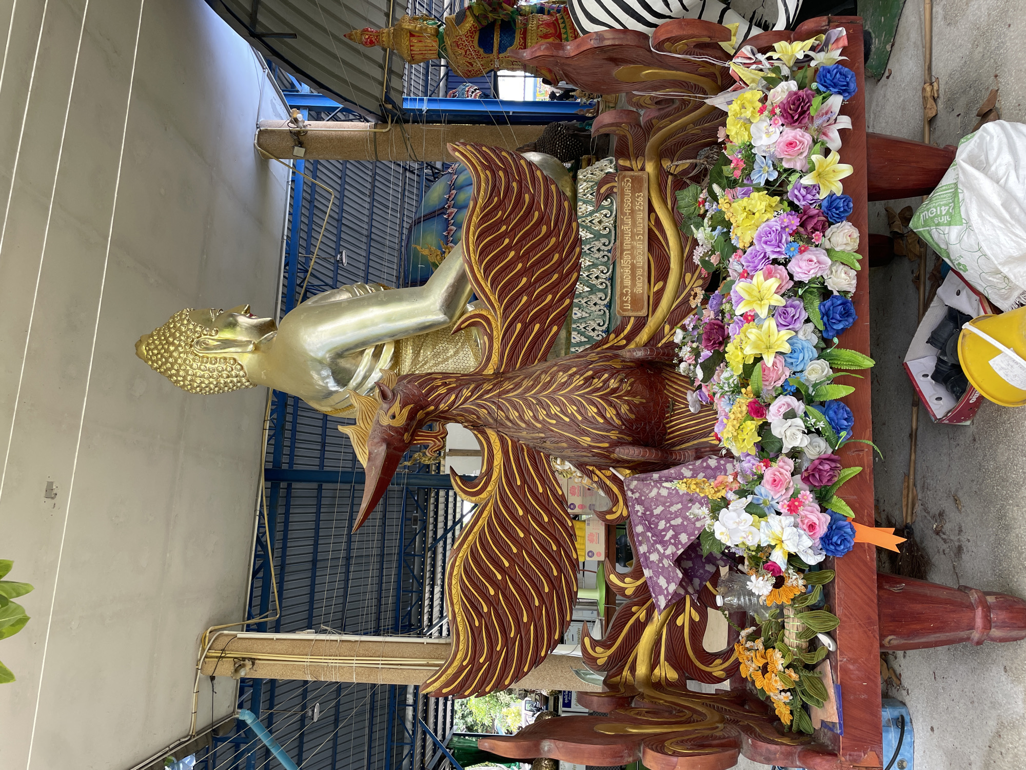 The image depicts an outdoor shrine in Bang Sare, Thailand, featuring a large golden Buddha statue in the background. In the foreground, a wooden platform is adorned with a vibrant display of artificial flowers in various colors, including pink, blue, yellow, purple, and white. A carved wooden bird, with intricate red and gold detailing, stands prominently on the platform, facing left. Behind the bird, a decorative wooden structure with red and gold wing-like extensions is visible. A small plaque with Thai script is attached to the base of this structure. To the right of the Buddha statue, another large, colorful statue, possibly a guardian figure, is partially visible. The environment appears to be under a covered structure with a blue metal roof and exposed support beams, suggesting it is part of an outdoor temple or shrine complex. The ground is concrete, with some debris, including plastic bags and a yellow bucket, scattered around the platform.
