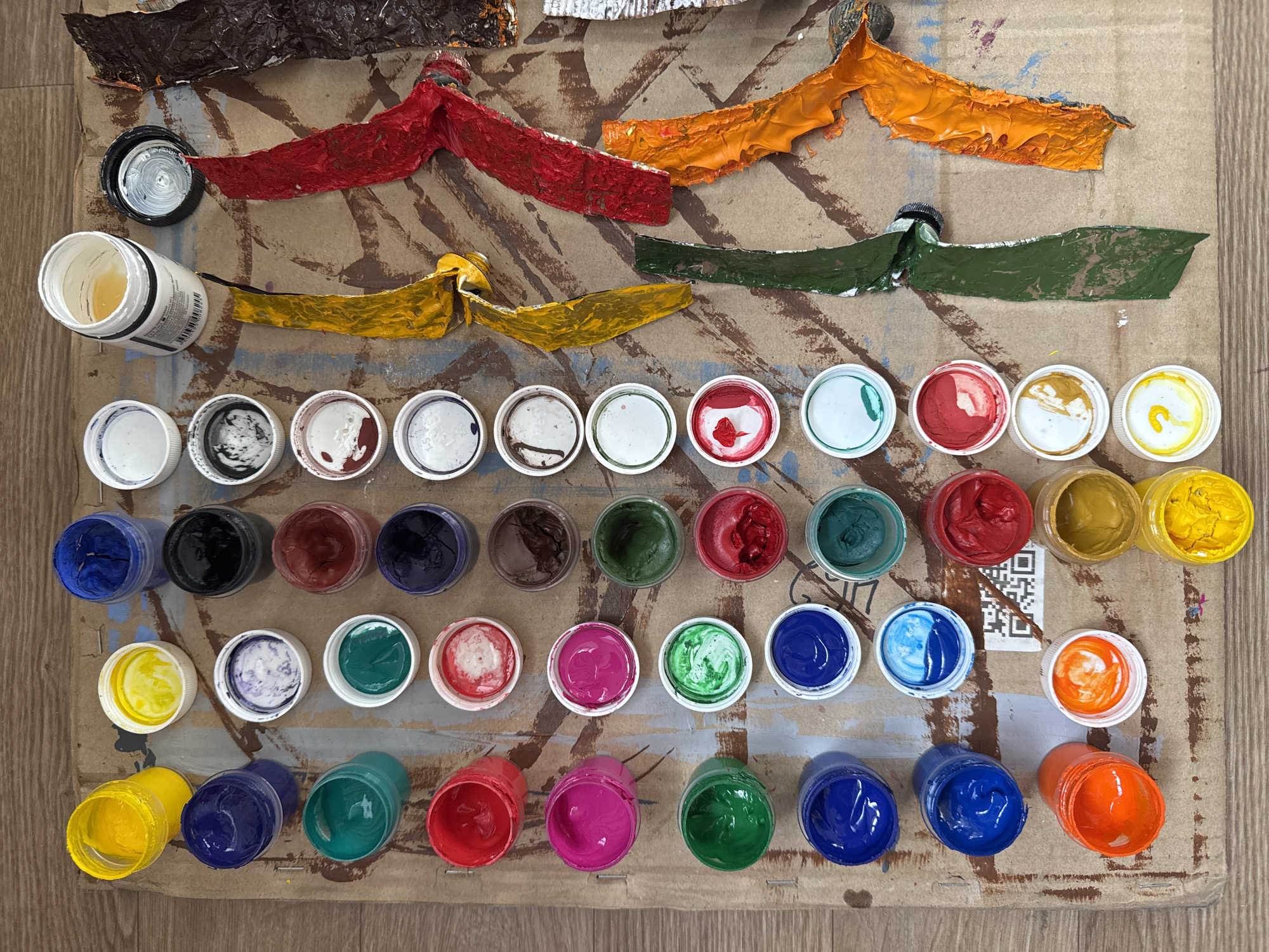 An overhead shot reveals a vibrant and messy art workspace, featuring an extensive collection of paints. The setting is indoors, with a heavily paint-splattered cardboard or textured surface, possibly resting on a wooden floor. The provided location context for this media file is Moscow, Russia.
At the top of the frame, several heavily squeezed and crumpled metal paint tubes are laid out horizontally. These tubes have dispensed thick, textured streaks of paint in colors including dark brown, red, orange, yellow, and dark green onto the surface.
Below these tubes, the main focus shifts to three organized rows of numerous small, open plastic pots or jars, each filled with liquid or gel paints. This diverse palette includes various shades of white, black, blue, green, red, yellow, brown, pink, purple, and orange. Many of the corresponding caps are placed beside their pots, also stained with residual paint. A larger white plastic jar, containing a yellow liquid or medium, is visible on the left, bearing a white label with small, illegible black text, though the word "PRODUCT" is partially discernible. Towards the center-right of the image, a black QR code is printed directly onto the cardboard surface.
The overall impression is one of active creative work, with a wide array of paints ready for use or having been recently used, evident from the diverse color selection and the heavily marked, expressive surface.
