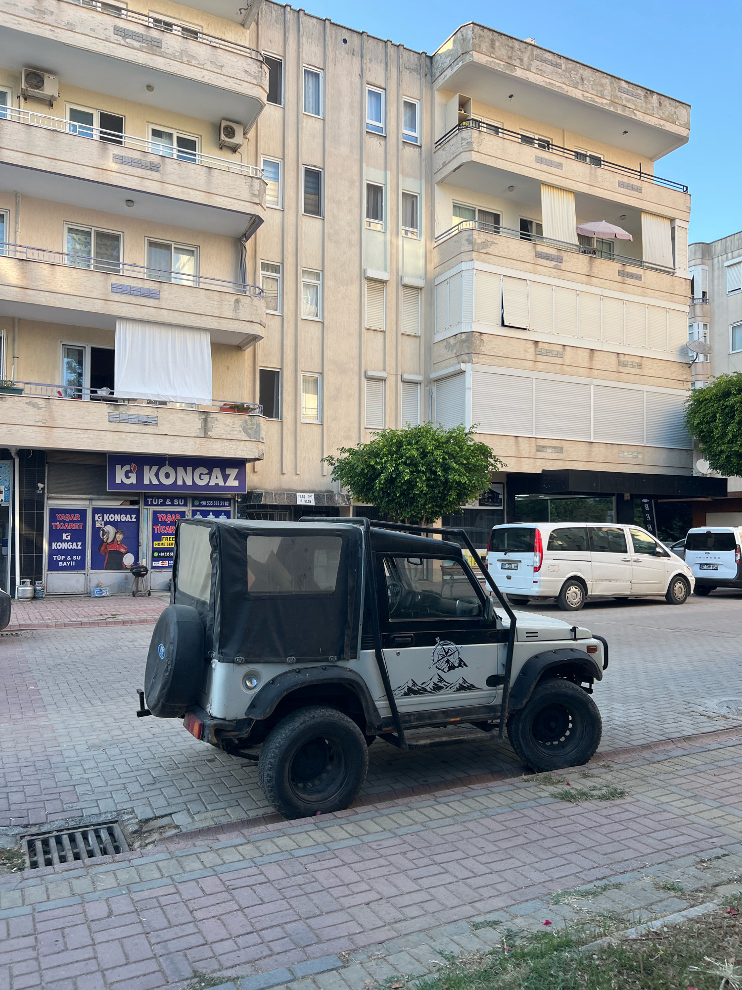 In Alanya, Turkey, a white four-door convertible SUV with a black soft top is parked on a paved street in front of a multi-story apartment building. The SUV has a spare tire mounted on the rear. The building's facade is light beige with balconies on each floor. Below the balconies, there are several shops, including one with a prominent "IG KONGAZ" sign. A white van and another white vehicle are parked further down the street. The sky is clear and blue. The street is paved with red and gray bricks, and there is a small patch of grass and a drain grate visible. The scene appears to be in a residential or commercial area of the city.