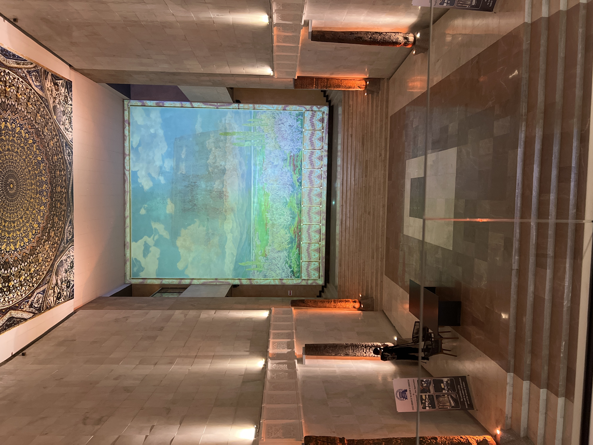 The image captures a grand interior space with a large mural or projection dominating the far wall. The mural depicts a landscape with a vibrant sky, cacti, and vegetation. The walls flanking the mural are made of large, beige stone blocks, illuminated by spotlights. The ceiling features an ornate, circular mosaic. In the foreground, there are marble floors and a set of tiered stairs. A person wearing dark clothing is standing on the left side, seemingly operating a camera or tripod, facing towards the mural. To their left, a signpost with images and text is visible. Two dark tables and chairs are situated in the middle ground on the left. The overall impression is one of a museum or cultural institution.