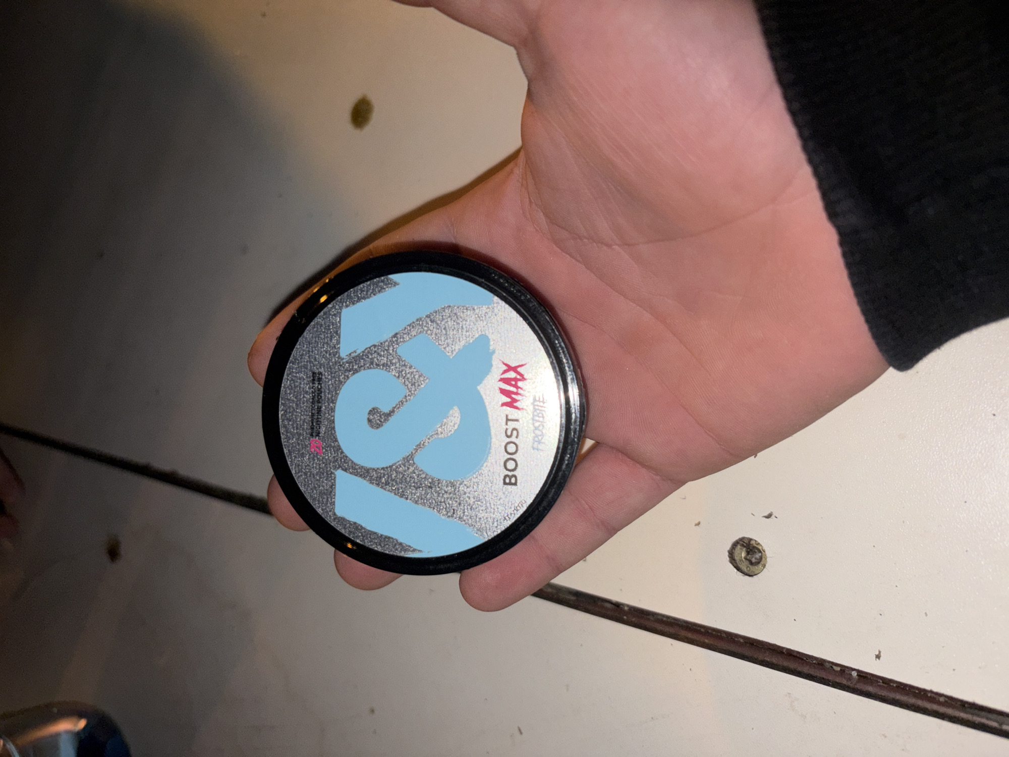 A close-up shot depicts a hand holding a round container with the words "BOOST MAX" and "FROSTBITE" on its label. The label features a stylized "19" in a light blue color. The hand is partially visible, showing skin tone. Part of a black sleeve is visible on the upper right. The container sits on what appears to be a white surface. There is no indication of the location being Plovdiv, Bulgaria.