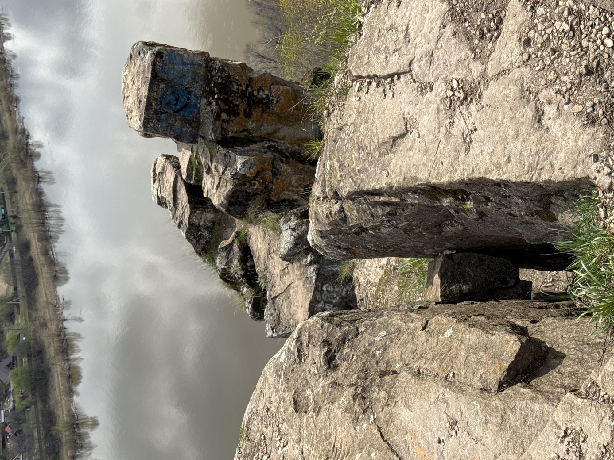 This image captures a rugged, natural landscape in Pavlovka, Russia, seen from an elevated, rocky vantage point overlooking a wide body of water, likely a river or reservoir.
In the foreground and midground, several large, weathered rock formations dominate the scene. These rocks exhibit varied textures, cracks, and shades of grey and brown, with patches of green grass and some lichen growing in crevices and on their surfaces. Small pebbles are scattered on top of some of the flatter rock surfaces. One prominent rock formation in the mid-distance features distinct blue graffiti, appearing as a stylized circular design or a smiley face. Another large rock in the immediate foreground acts as a natural step or ledge.
The background is dominated by the calm, grey surface of the water, which reflects the overcast sky, indicating daylight hours on a cloudy or hazy day. Along the far bank of the water, a narrow strip of land is visible, lined with trees that are mostly bare or showing very early signs of spring foliage. Further in the distance, small structures, likely houses or buildings, can be faintly discerned nestled amongst the trees.
No specific activity or event is taking place; the image presents a serene, static landscape view. The overall impression is one of natural beauty and rugged terrain typical of a riverside or lakeside cliffside.