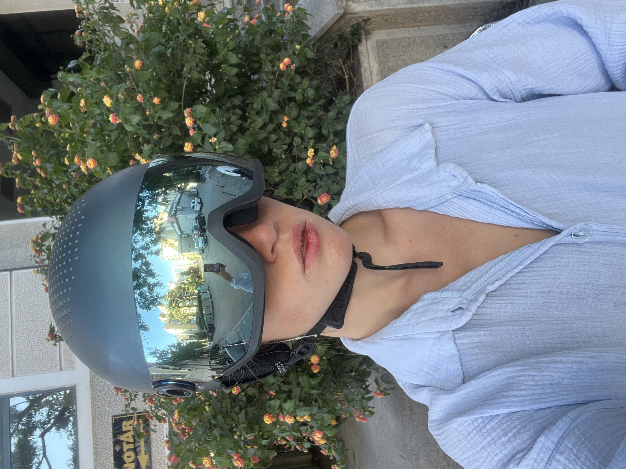 A woman wearing a blue button-down shirt and a dark helmet with reflective visor, is positioned in front of a building near an office. The helmet's visor reflects the cityscape of Budva, Montenegro. The woman has light skin, red lips, and a black strap securing the helmet. The background includes flowering bushes and a building with the word "NOTAR" on a sign with an arrow pointing to the left.