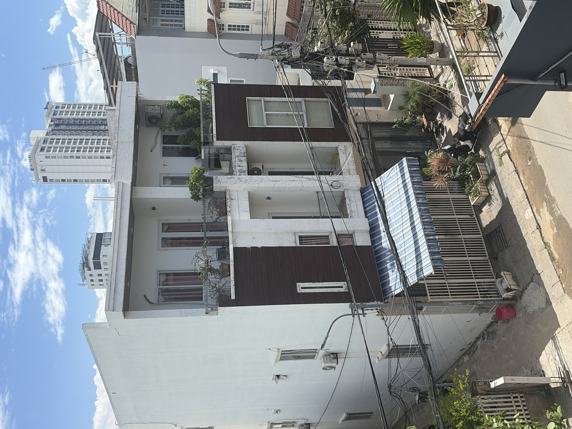 The image shows a narrow street in the city of Đà Nẵng, Vietnam, lined with multi-story buildings. The foreground is dominated by a white, three-story building with a dark brown facade on the second story. It features multiple windows, balconies with plants, and an air conditioning unit. A striped awning covers a portion of the ground floor entrance, which is secured by a white metal fence.
Behind the foreground building, a taller, modern white skyscraper with many windows stands prominently, identified by the "HIYORI" logo at the top. A construction crane is visible in the distance. Other buildings in the scene include a multi-story structure with a red tiled roof and several other residential or commercial buildings with various architectural styles.
The street is paved and appears somewhat weathered, with some areas of dampness. Numerous electrical wires are strung across poles and buildings, indicating the urban setting. Several potted plants and some small trees are visible in front of the buildings and along the street. A black scooter is parked on the side of the road. The sky is blue with scattered white clouds.