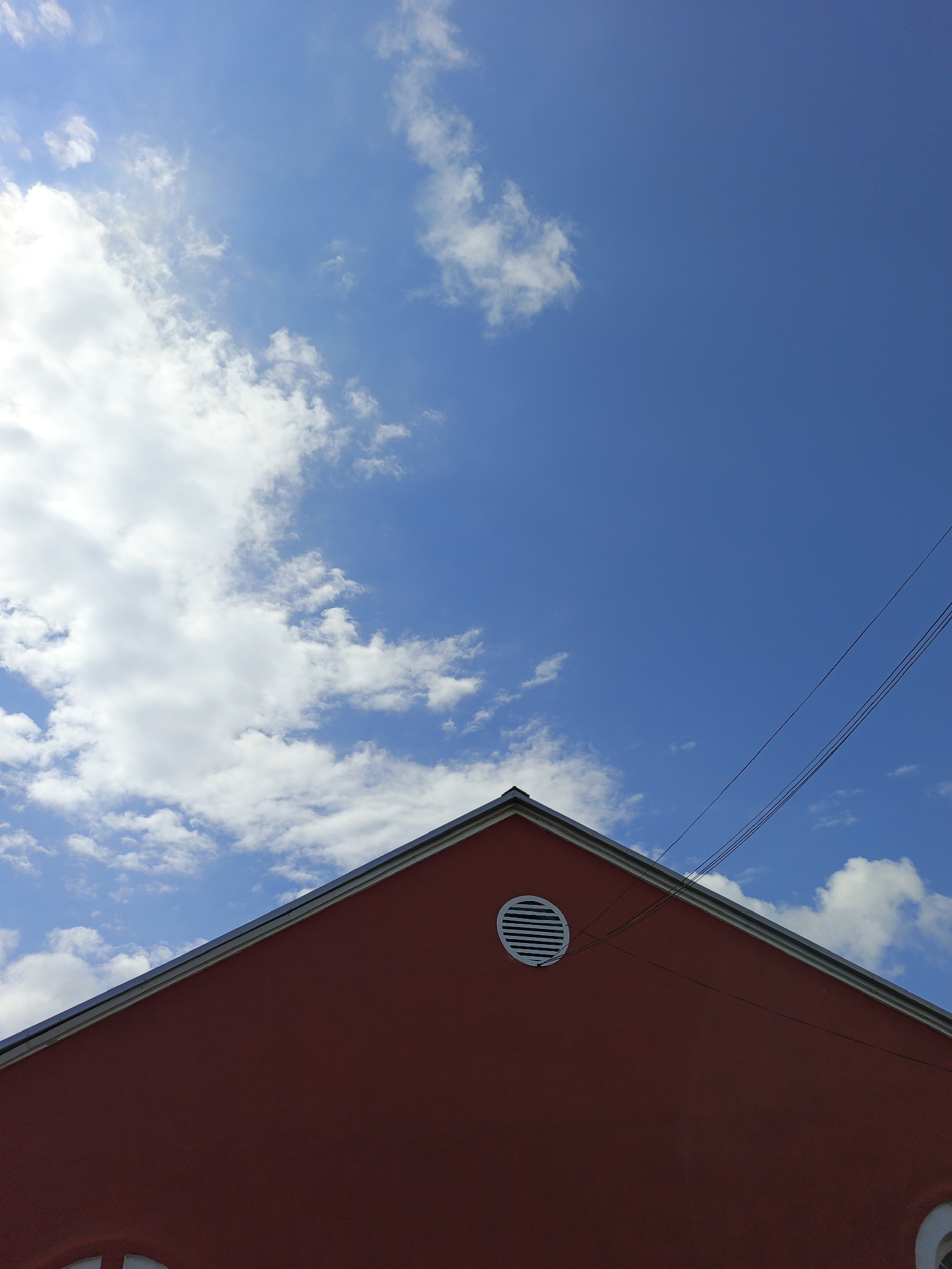 The image is a low-angle shot of a red-colored building's upper portion against a bright blue sky with scattered white clouds. The building has a peaked roofline with a grey trim. A round, white vent with horizontal slats is visible on the facade. Three dark electrical wires run diagonally across the sky, originating from the right side of the frame and extending towards the upper left. The scene is devoid of people or any discernible activity, presenting a still and architectural view. This image was captured in Medvedyovka, Russia.