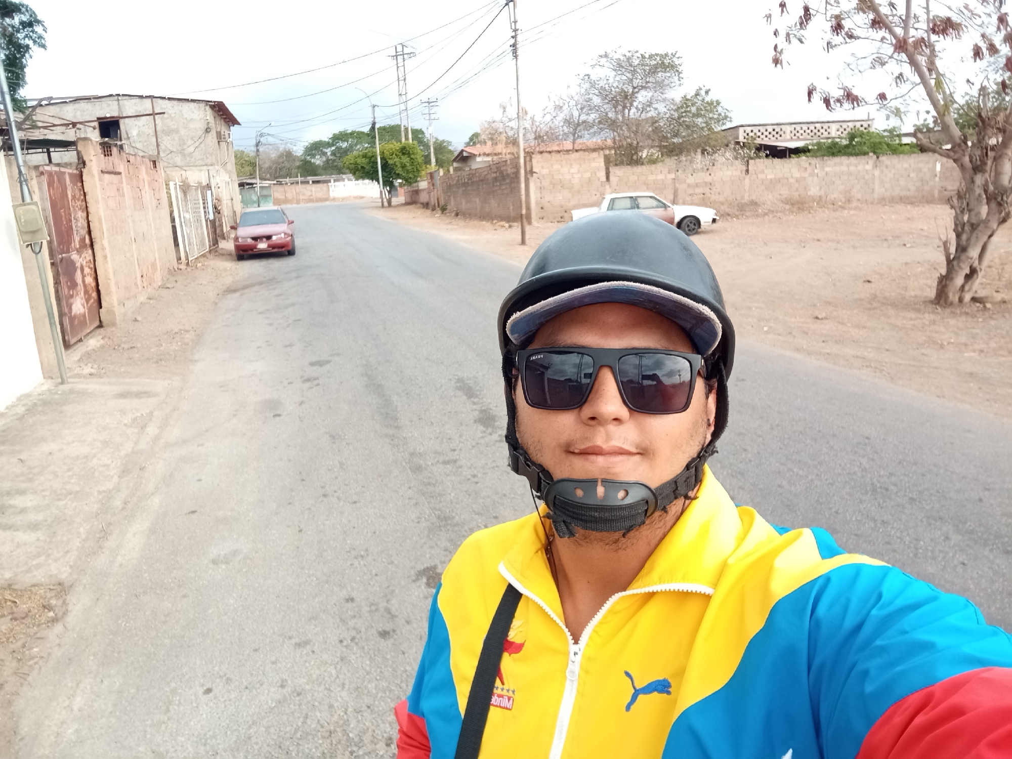 A man wearing a black helmet and sunglasses stands on a paved road in Juan Griego, Venezuela. He wears a yellow, blue, and red jacket. Behind him is a road with houses and buildings on either side. A red car is parked on the left side of the road. Electrical wires are visible above the street. A light-colored pickup truck is visible further down the road on the right. A leafless tree is also on the right side of the road.