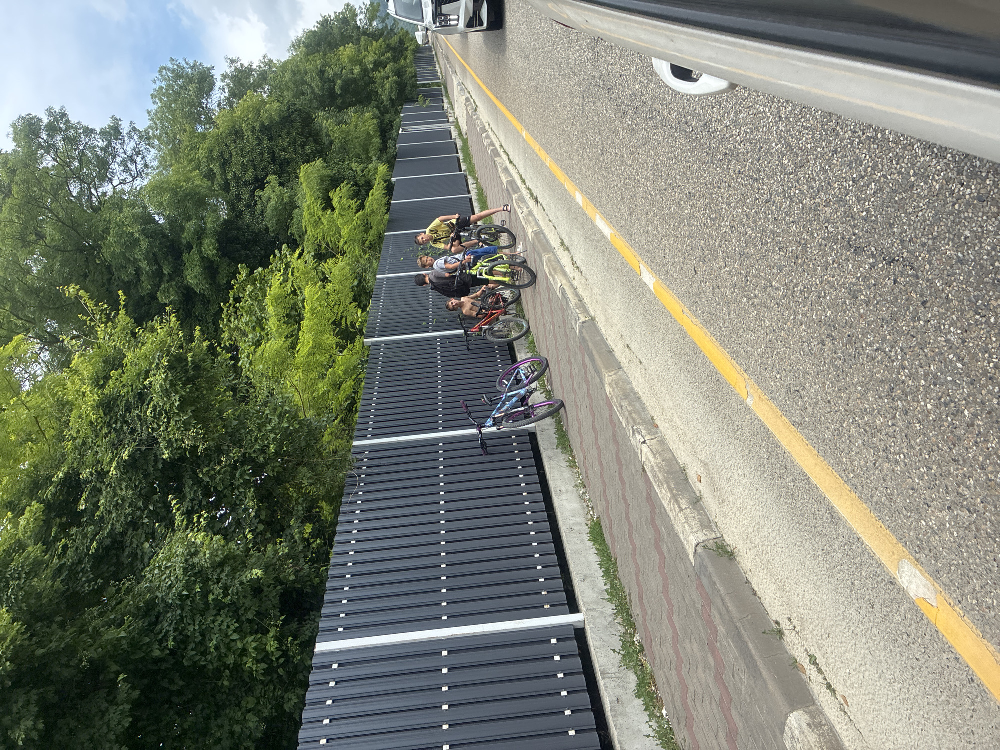 Four young males are gathered with bicycles on a paved sidewalk beside a dark, corrugated metal fence. The males are positioned between bicycles. One male is sitting on a red bicycle, facing away from the camera. Another male is standing beside a bright green bicycle. A third male is standing next to the green bicycle, also facing away from the camera. A fourth male, wearing a yellow shirt and dark shorts, is standing with a black bicycle. The sidewalk is bordered by a concrete curb and a strip of gravel with sparse vegetation. Beyond the fence, dense green foliage is visible, suggesting a wooded area. The scene appears to be on the side of a paved road, indicated by a yellow line marking the lane. A white vehicle is partially visible on the right side of the road. The sky is cloudy. The location is identified as Arkhipo-Osipovka, Russia.