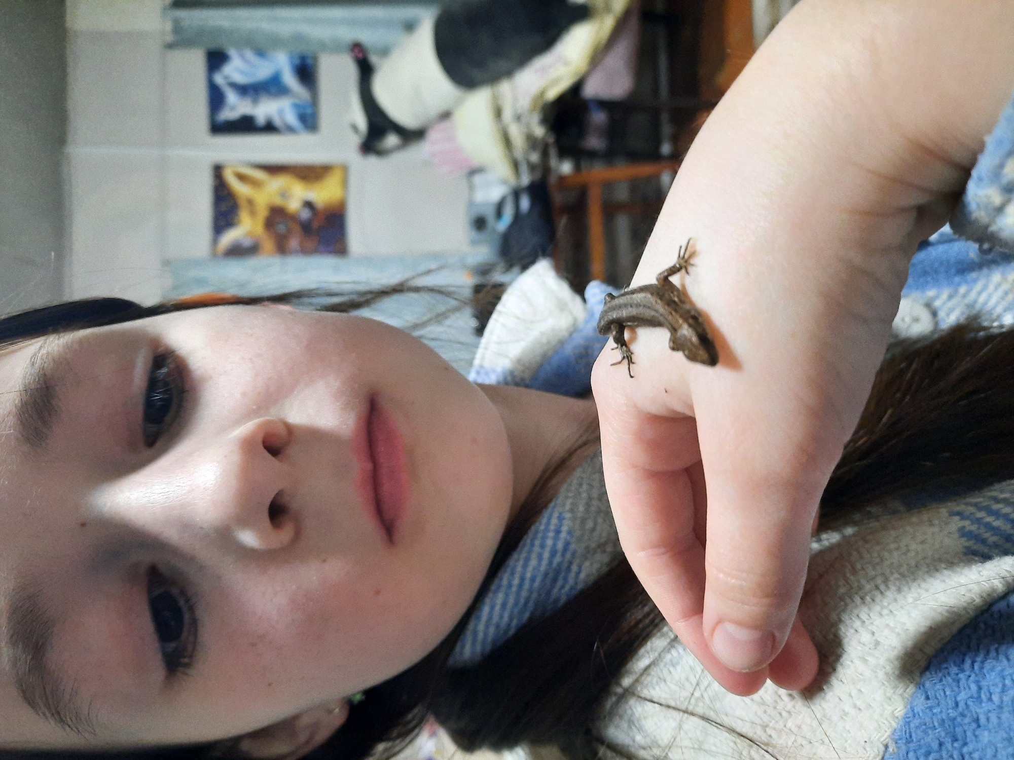 A young person is lying down, looking at the camera. A small lizard is on their hand. The surrounding environment includes what appears to be a room with pictures on the wall and various items scattered around. There is no verifiable indication in the image to determine this photo was taken in the city of Krasnogorskoye, Russia.