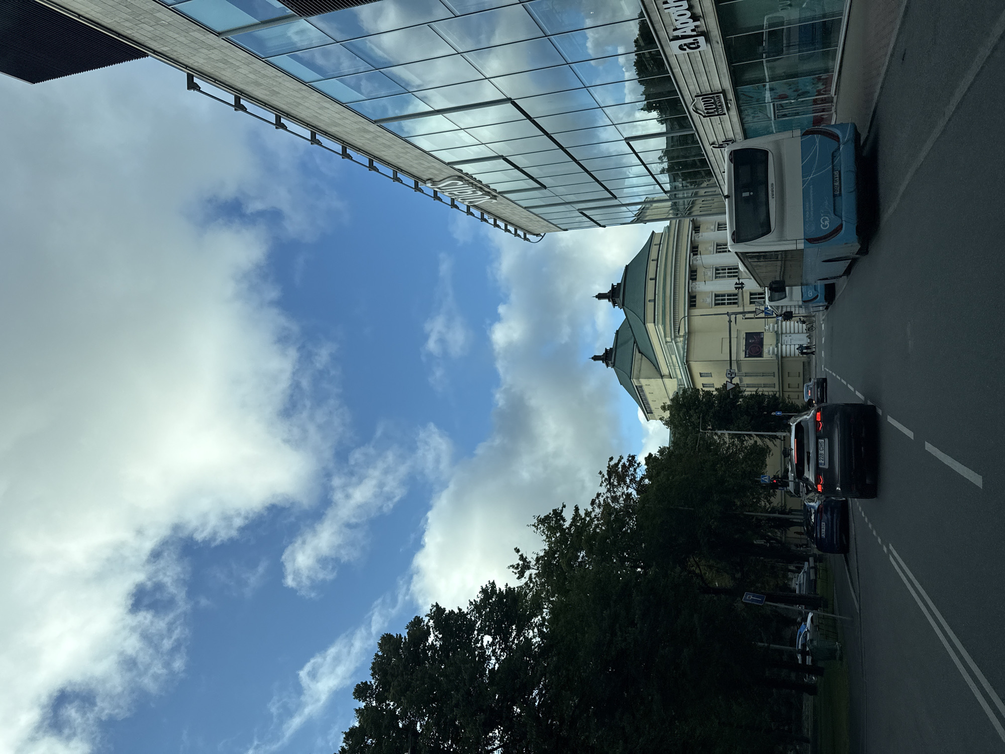 The image depicts a street scene in Tallinn, Estonia, likely in the city center given the presence of modern architecture juxtaposed with a grand historical building. In the foreground, a dark-colored SUV is driving down a road with white lane markings. Behind it, a light blue and white bus is parked on the right side of the road. To the right of the bus, a modern building with large glass windows and the name "SOLARIS" visible at the top stands. Reflections of the sky and trees are visible in the glass. Further in the background, a yellow building with a green roof and domed structures is prominent, identifiable as the Estonia Theatre. Lush green trees line the left side of the road. The sky is blue with scattered white clouds. A few pedestrians are visible on the sidewalk near the historical building. The overall impression is a contemporary urban environment with a blend of old and new architecture.
