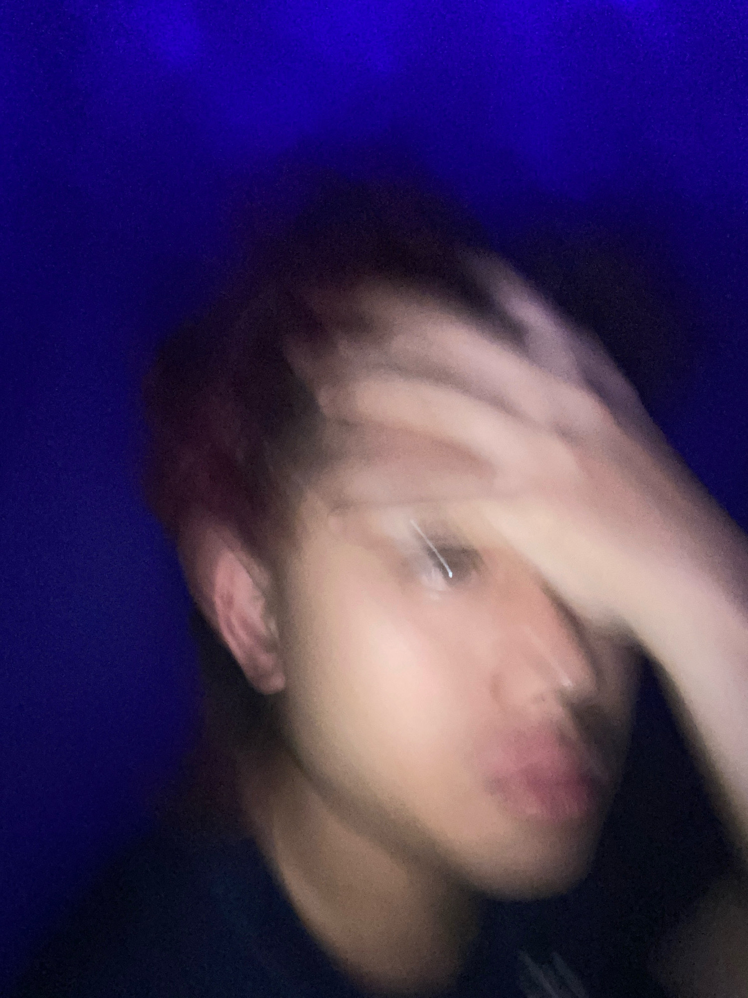 The image is a blurry, close-up portrait of a young person with reddish-brown hair. The person's right hand is raised and obscures part of their face. A bright blue background fills the upper portion of the image. The location in Klurak, Indonesia is not discernible from the image.