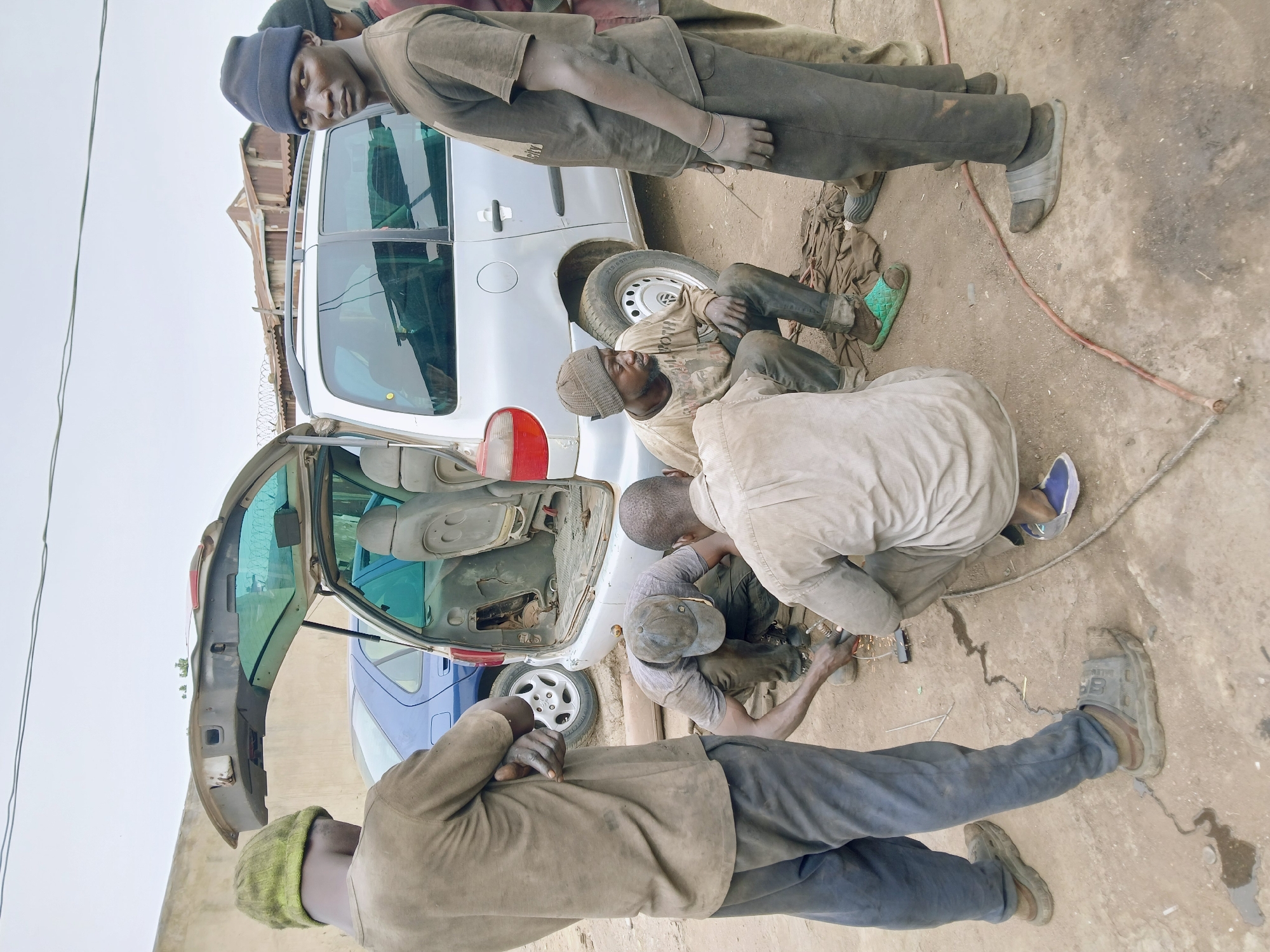 The image captures an outdoor auto repair scene in Rafin Par, Nigeria, featuring five men engaged in vehicle maintenance. The setting appears to be an informal workshop or roadside area with a dirt and concrete ground.
In the foreground, a man wearing a light brown shirt, dark trousers, and a lime-green beanie stands with his back largely to the viewer, observing the work. To his right, another man, also in work-worn clothes and a dark blue beanie, stands near a silver hatchback car and looks directly at the camera with a neutral expression. This silver car, possibly a Renault Clio, has its rear hatch open, exposing its interior, and appears to be the primary focus of the repairs. Its rear passenger-side tire, showing some wear, is visible.
Three other men are actively working on the silver car. One man is seated on the ground near the rear of the silver car, leaning against it, dressed in a striped beanie and a light-colored, soiled t-shirt. Further right, two men are crouched or kneeling, attending to the open trunk area of the silver car. One of these men wears a dark cap and appears to be operating a tool that generates sparks, suggesting welding or grinding work.
A blue car is partially visible behind the silver vehicle. The ground is dusty and uneven, with a reddish cord or hose running across it. The lighting indicates daytime conditions, likely overcast, as there are no distinct shadows. The men's clothing is uniformly soiled and practical, consistent with manual labor. No discernible text is clearly visible on any objects or clothing within the frame.