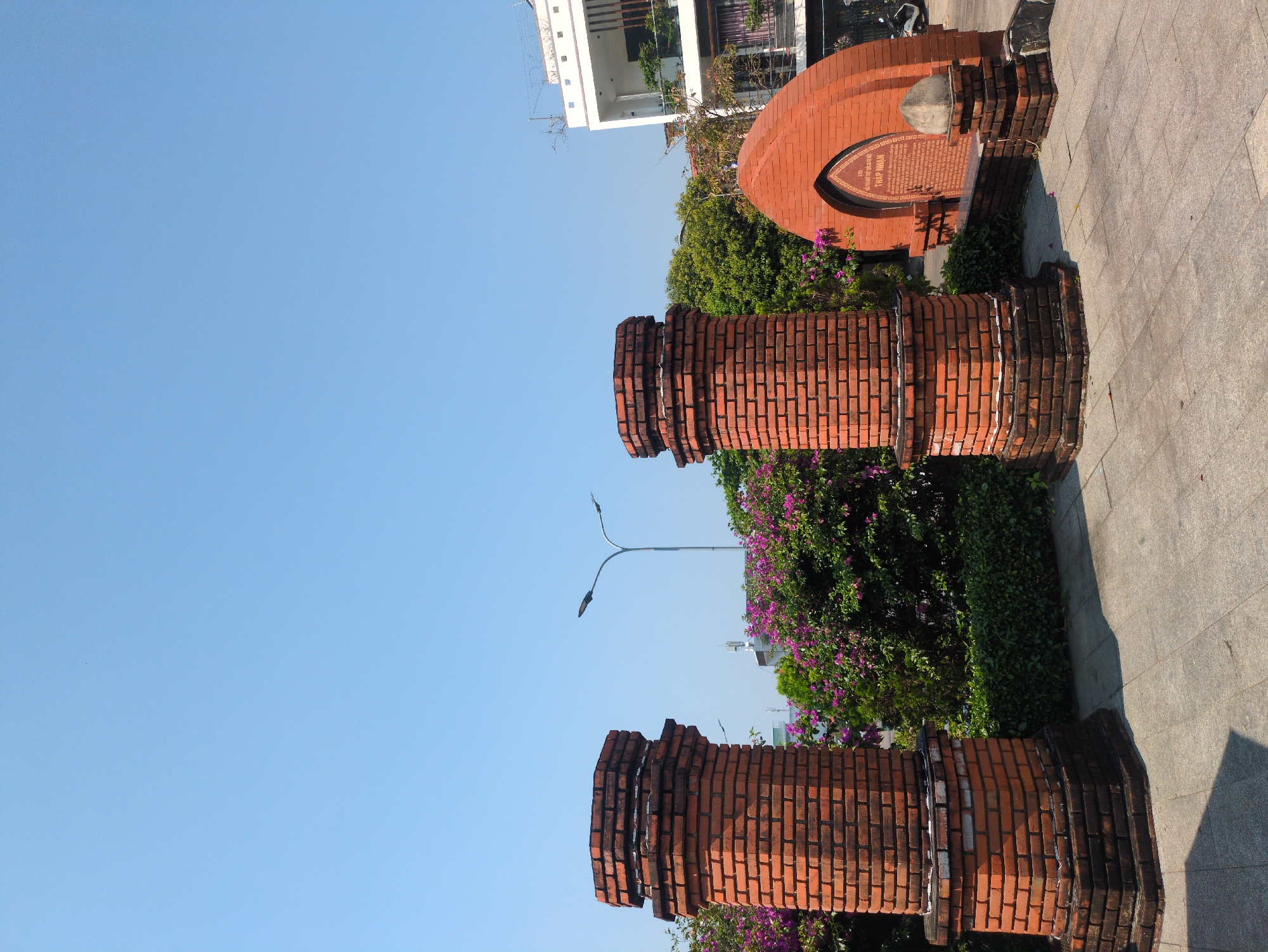 The image depicts a brick structure monument in Tuy Hòa, Vietnam. The foreground features two brick pillars with ornamental tops and bases, surrounded by green shrubs and purple flowers. A street lamp post rises behind the pillars. In the background, a semi-circular brick structure with a plaque is visible. A paved walkway runs along the side. A white building is present in the background under a clear blue sky.