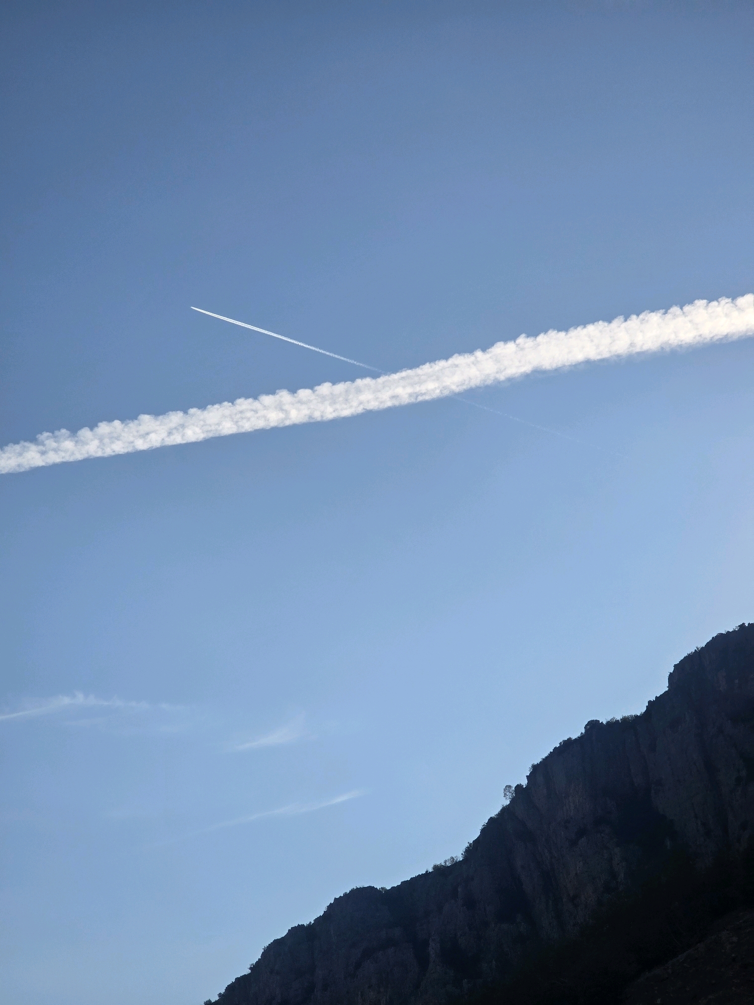 This media file captures a dramatic sky over a mountainous landscape. The sky is a clear, vibrant blue, suggesting a bright, sunny day. Two prominent contrails streak across the sky, indicating the passage of aircraft at high altitude. The lower portion of the image is dominated by the dark, rugged silhouette of a mountain range. The texture of the mountain suggests steep, rocky slopes with sparse vegetation clinging to its surface. The contrast between the vast, open sky and the solid, imposing mountain creates a sense of scale and natural grandeur. The lighting suggests it might be late afternoon or early morning, as the mountain is casting a shadow and the sky has a warm tone. The overall mood is one of vastness and natural beauty, with a hint of human presence suggested by the airplane trails. The setting appears to be a remote, mountainous region, fitting for the provided location context of Podgorica, Montenegro. There is no visible text in the image.