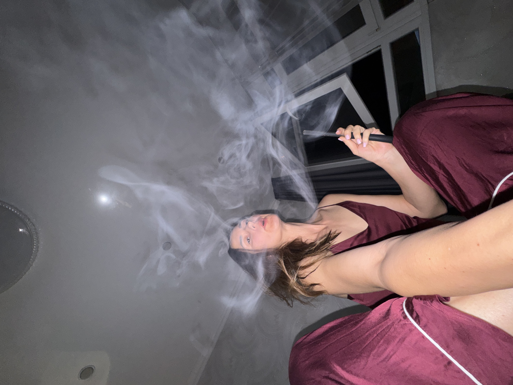 The image depicts a female individual indoors, captured from a low-angle perspective, likely a selfie. The woman, an adult with long brown hair, is wearing burgundy-colored loungewear consisting of a camisole top and pants with white piping. She is seated and observed exhaling a large cloud of white vapor or smoke from her mouth, while holding a slim, dark cylindrical object, consistent with a vape pen or electronic cigarette, in her right hand. Her fingernails are painted pink.
The surrounding environment is a dimly lit room with grey, textured walls and a light-colored ceiling. Recessed lighting fixtures and a prominent circular fixture are visible on the ceiling. In the background, to the right, are large, multi-paned windows, one of which is partially open, indicating an outdoor nighttime scene. Dark curtains are partially visible adjacent to the windows. The overall lighting is subdued, with reflections visible on the ceiling and from the windows. The location is described as being in the city of Sochi, Russia, although this specific geographical detail is not visually discernible within the image itself.