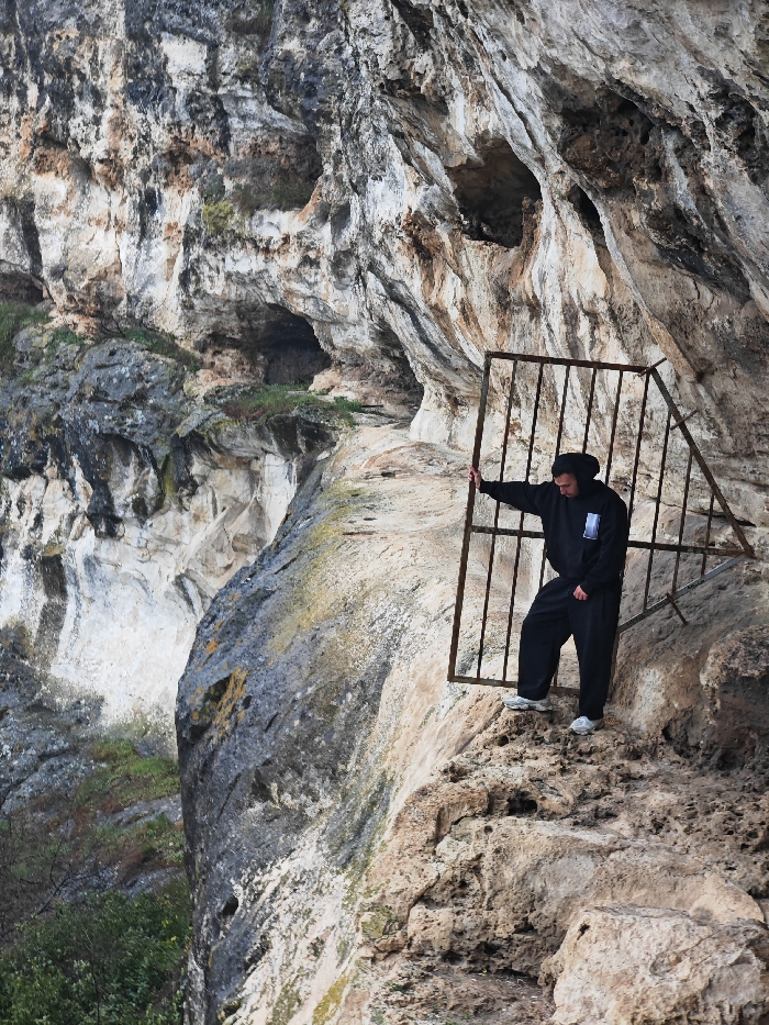 A male individual is observed standing on a natural rock ledge in the Shumen Plateau, Shumen, Bulgaria. The subject is dressed in a black hooded sweatshirt, black loose-fitting trousers, and white athletic shoes. Their left hand rests on a vertical bar of a rusted, dark-colored metal grate structure positioned behind and to their right, leaning against the prominent limestone cliff face. The cliff displays varied stratification, multiple natural cave openings, and colorations ranging from white and light grey to tan and dark grey. The rock ledge to the immediate left of the subject descends steeply. Sparse green vegetation is visible in crevices along the lower sections of the cliff. The scene is illuminated by ambient daylight.