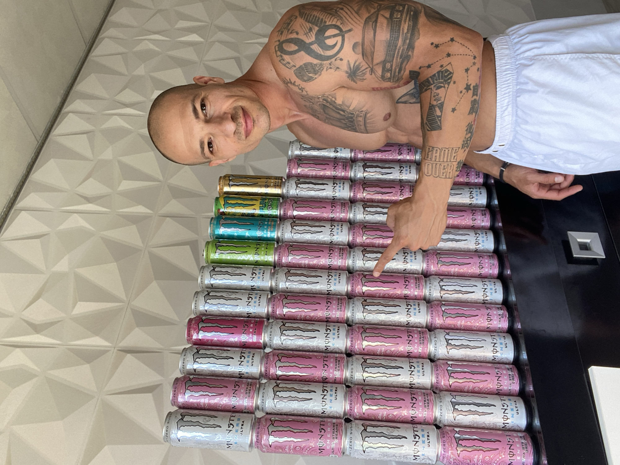 A shirtless, muscular man with a shaved head and tattoos on his arm and torso stands in front of a wall of stacked Monster Energy cans. He is wearing white shorts and is pointing to the cans with his right hand. The cans are arranged in rows and columns, with different flavors visible, including pink, silver, green, blue, and gold. The wall behind the cans has a textured, geometric pattern. The lighting suggests an indoor environment with natural light. The man is smiling and looking towards the camera. The scene appears to be set up for a photo opportunity. There is no discernible indication of the city of Pleven, Bulgaria.