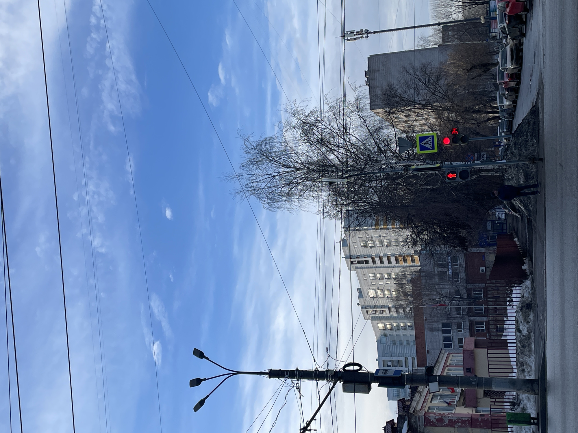 The image displays an urban street scene in Izhevsk, Russia, specifically along Ulitsa Svobody near its intersection with Kommunarov Street, facing southeast. The sky is a bright blue with scattered thin white clouds, crisscrossed by numerous overhead utility lines.
In the left foreground, a weathered two-story building (Ulitsa Svobody 175) with a faded yellow-pink facade and a red roof displays two small Russian flags. A dark metal fence stands in front of this building, with irregular piles of melting snow and a green waste bin adjacent to the curb. A utility pole with a three-lamp street light, extensive cabling, and a blue-and-white bus stop sign is positioned centrally on the left.
The street surface in the immediate foreground is wet. A single pedestrian, dressed in dark clothing, stands on the sidewalk facing away from the camera, positioned next to a traffic light pole. The pedestrian signal displays a red standing man icon, and the vehicular traffic light below it is red with a digital countdown showing "34". A blue-and-white square sign indicating a pedestrian crossing is mounted above these lights.
On the right side of the street, a line of parked passenger vehicles is visible. Patches of slushy snow are accumulated along the curbs and sidewalks. Bare deciduous trees, indicating a dormant season, are distributed along the street, with one prominent tree in the center-right foreground.
In the midground, a gray brick two-story building (Ulitsa Svobody 175A) is visible behind the initial structures, featuring Cyrillic signage "ГАРМОНИЯ" (Harmony) and two small flags. Further into the background, several modern multi-story residential buildings, predominantly gray with reflective windows, rise above the cityscape. A large, older, monolithic gray building is visible to the right, and a tall cellular communication tower stands on the far right. No active interactions or moving vehicles are present within the frame.