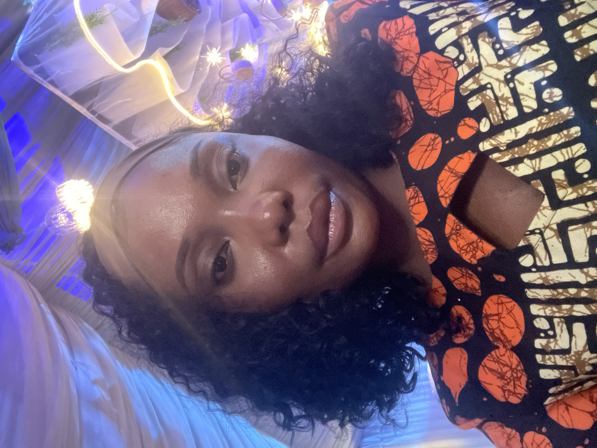 A close-up, slightly tilted selfie shows a Black woman with dark curly hair, wearing a black and orange patterned top and a light-colored headband. Her face is well-lit, with a slight smile. The background appears to be a decorated space with white fabric draping and string lights, creating a festive or celebratory ambiance. There is no verifiable indication in the image to confirm a location in Jos, Nigeria.