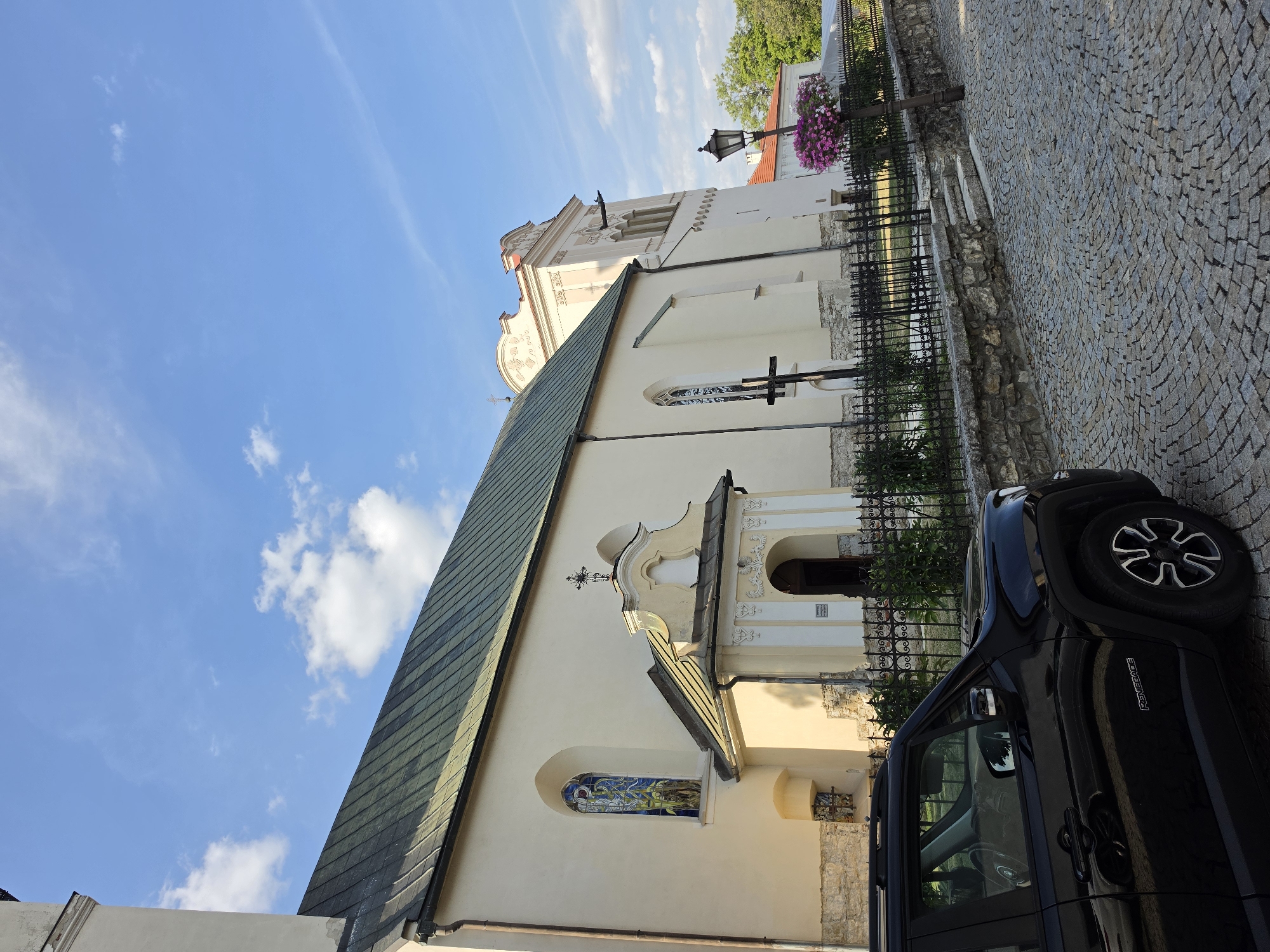 In an outdoor, urban environment, a black Jeep Renegade is parked on a cobblestone street in front of a cream-colored church. The church features a green-tiled roof and a large stained-glass window. A decorative entrance with a cross above it is visible, as is a black metal fence enclosing a garden area. A streetlamp with a hanging flower basket stands to the right of the church, and a small, weathered stone staircase leads up to the fence line. The sky is a clear blue with scattered white clouds. The image is captured from a low angle, looking up at the church and sky.