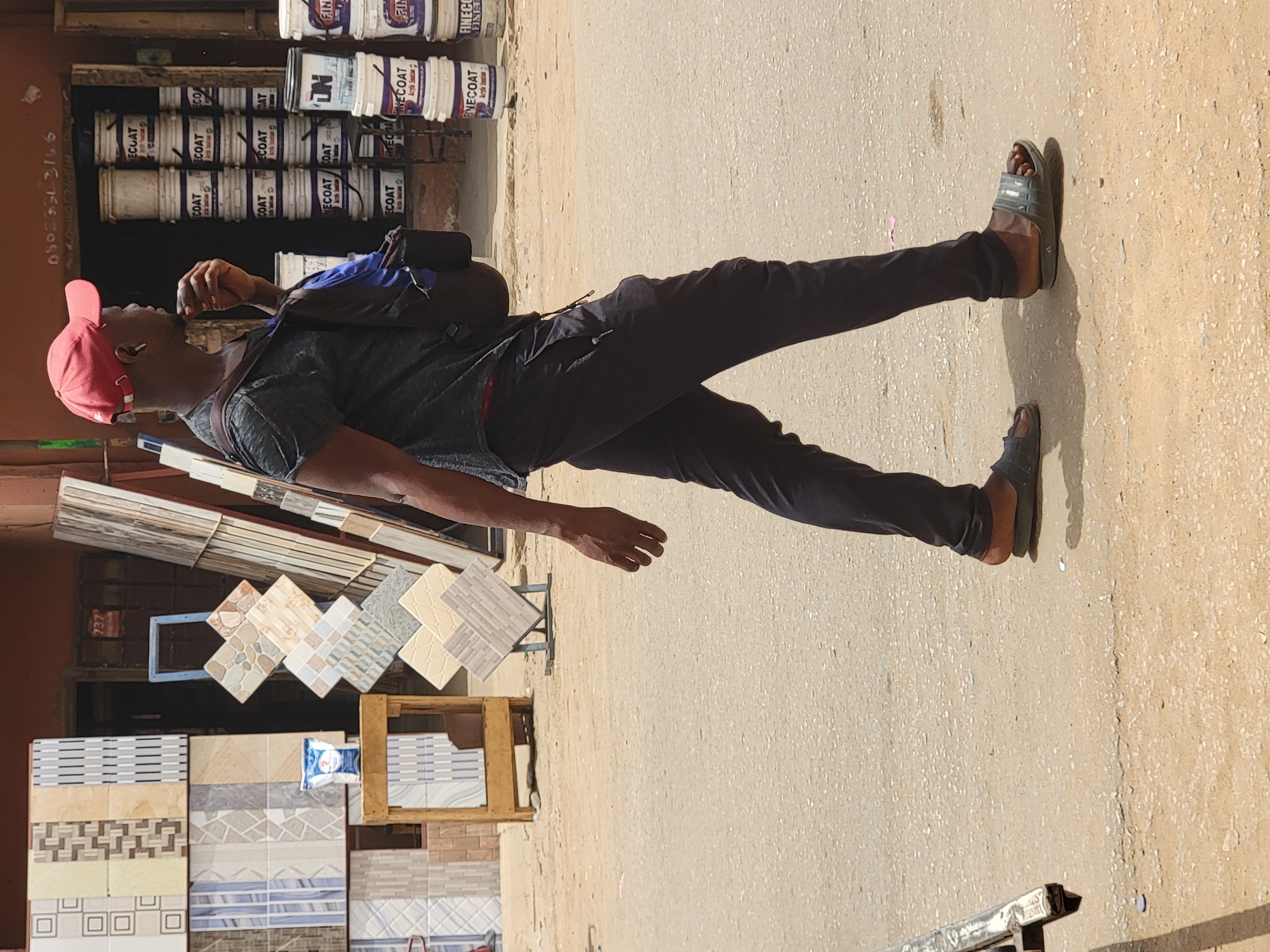 A man stands on a street in Maiburiji, Nigeria, next to a tile vendor and across from paint cans. The man wears a red cap, dark t-shirt, black pants, and sandals. He also has a backpack. Beside him, a display of various tiles is visible. Across the street, stacked paint cans are visible in a store. The pavement is visible, and the image is brightly lit. A phone number, "0802533166," is painted on a building.