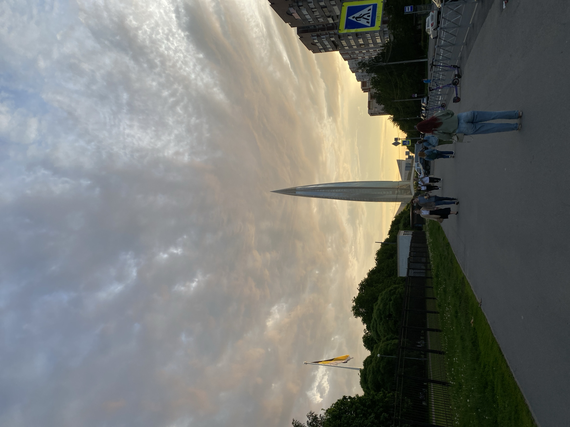 This image captures an urban landscape in Saint Petersburg, Russia, with the prominent Lakhta Center skyscraper towering majestically in the middle distance. The scene is illuminated by the soft, warm light of what appears to be late afternoon or early evening, with dramatic, partially overcast skies showcasing a mix of grey, white, and subtle golden hues where sunlight penetrates the clouds.
In the foreground, a paved pathway runs alongside a grassy strip and a line of trees. Several people are seen walking along this path, including a person with red hair wearing a green top and jeans, seen from behind in the mid-right. Further down the pathway, other pedestrians and a row of parked electric scooters indicate a lively urban setting. To the left of the pathway, a black fence borders an area of dense greenery, beyond which residential or commercial buildings are visible.
A blue pedestrian crossing sign, featuring a white walking figure, is prominently displayed near the top left corner, attached to a utility pole next to a building. Another notable object is a black and yellow striped flag, possibly the imperial flag of Russia, flying from a flagpole in the lower left section of the image, partially obscured by trees. The overall impression is one of everyday urban life unfolding against a striking natural backdrop and modern architecture.