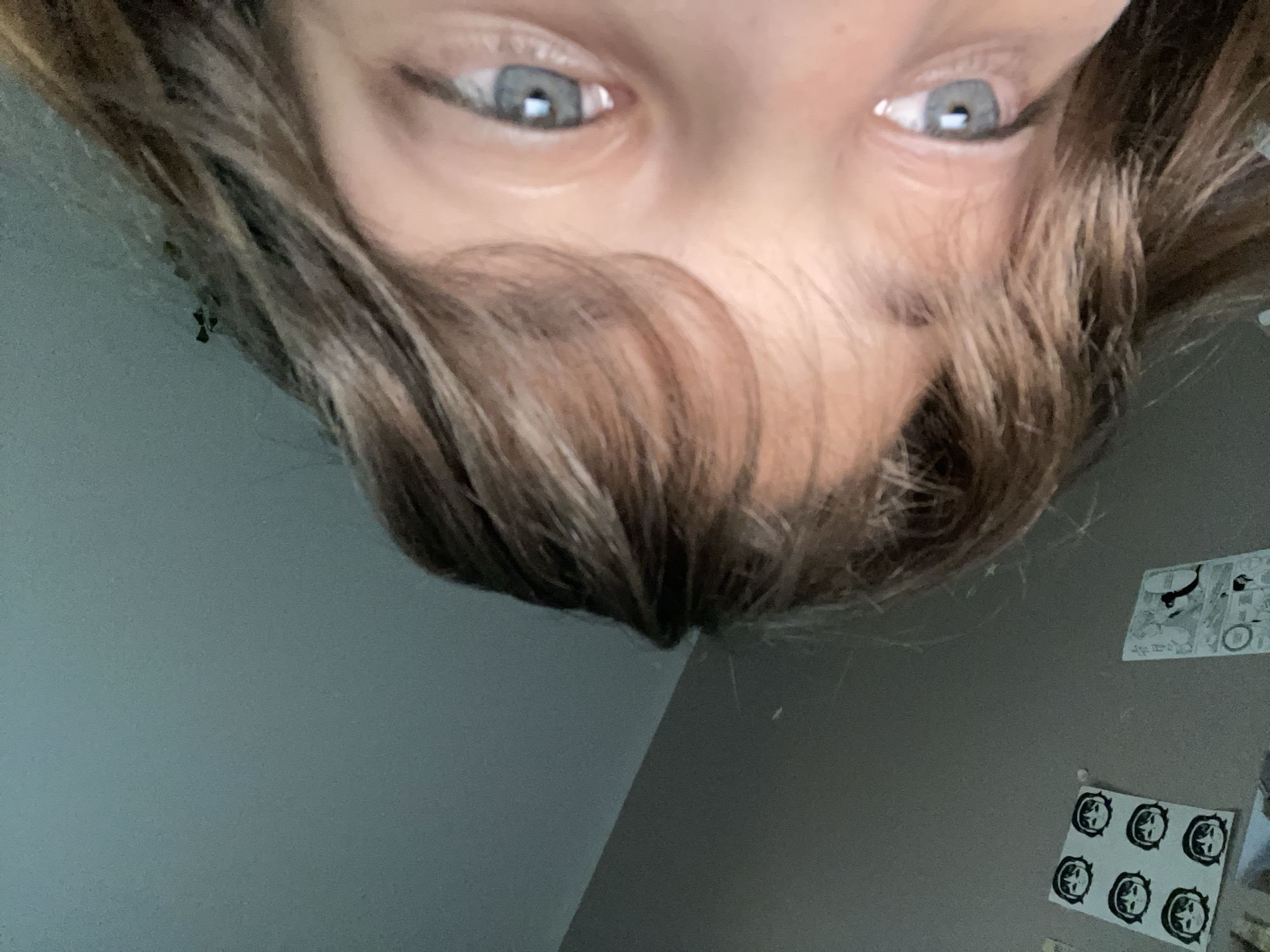 The image, taken from a low angle, shows a close-up of a person's face, focusing on their eyes and hair. The person has light-colored hair that partially covers their face, with strands falling across their forehead and cheek. Their eyes are light blue and appear to be looking upwards. The background is a plain, grey wall with some faint, decorative stickers or posters. On the left side of the wall, there is a cluster of six identical circular stickers featuring cartoon faces with black hair and eyes. To the right of these, there is a larger poster with black and white line drawings, possibly from a comic or manga. The lighting suggests an indoor environment, possibly a bedroom, with natural light coming from an unseen source. The image does not contain any information that would allow for its location to be determined in Мухавецкі сельскі Савет, Belarus, or any other specific location.